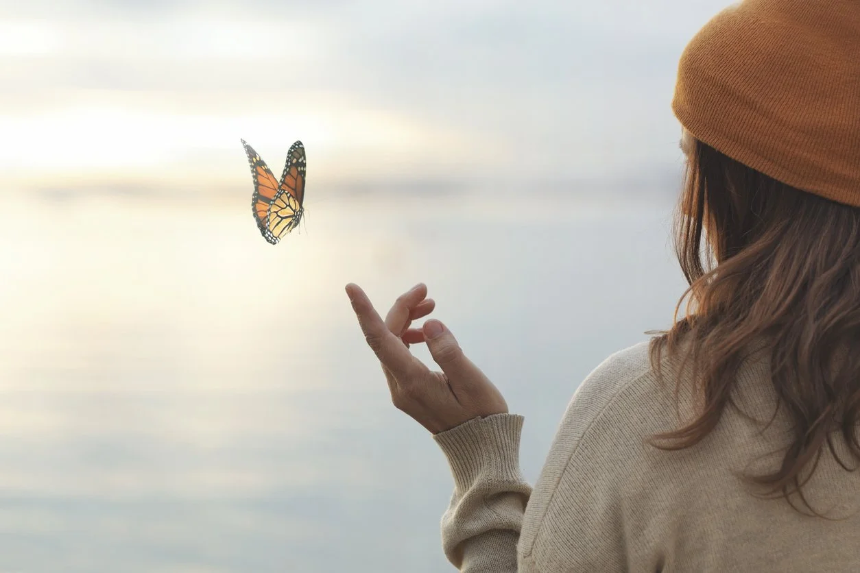 A woman with long hair wearing a brown beanie and beige sweater reaching out towards a Monarch butterfly near a body of water during a cloudy day.