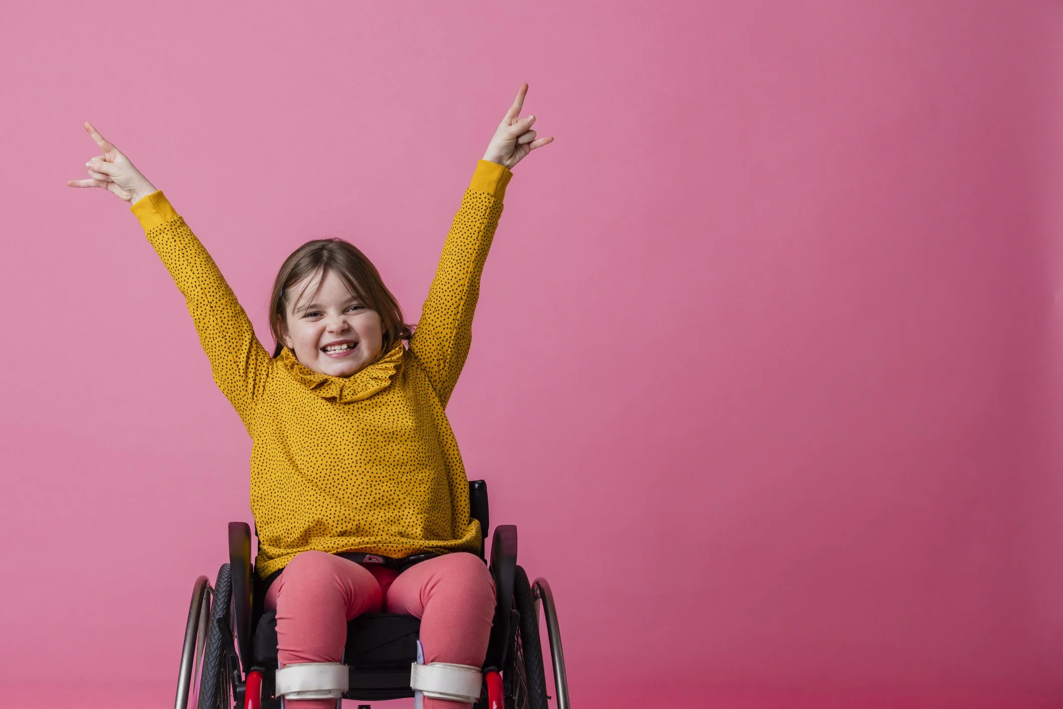 A young girl with brown hair and a bright smile wearing a yellow dotted long-sleeve shirt and pink pants, sitting in a wheelchair with a pink background. She has both arms raised with fingers pointing upwards.