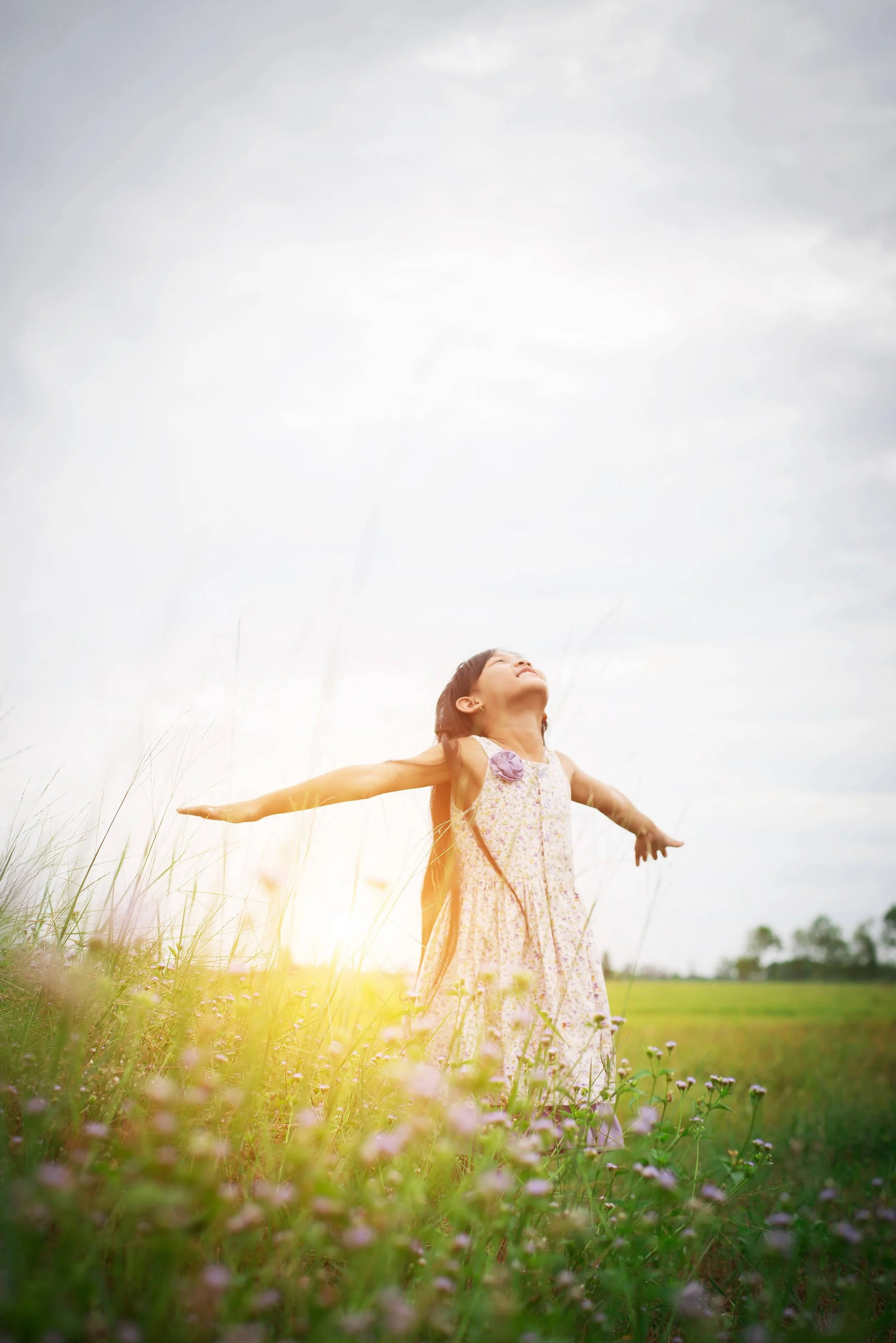 A young girl in a floral dress standing in a grassy field with her arms outstretched, looking up at the sky as the sun sets or rises.