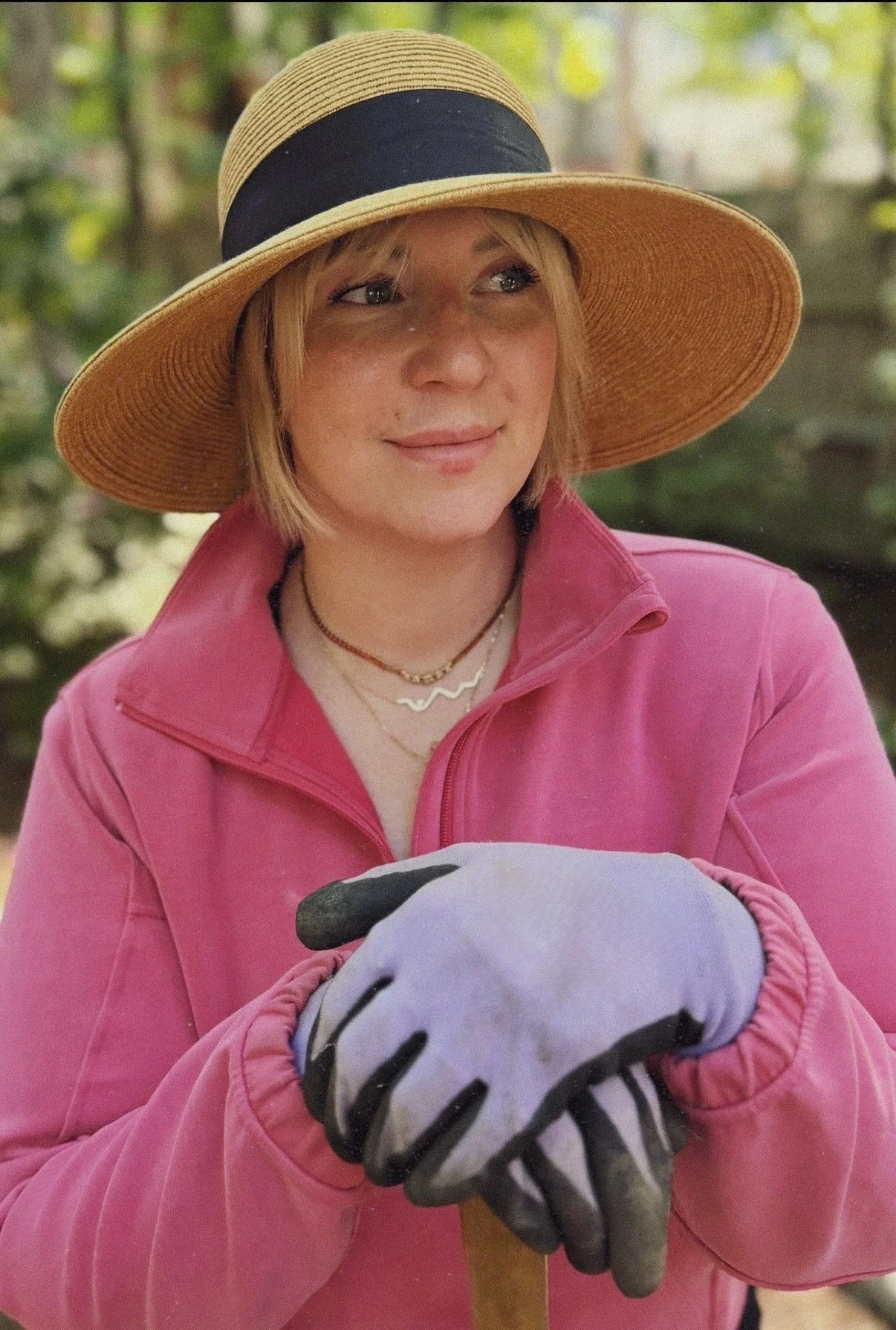 A woman wearing a large straw hat with a black band, a pink jacket, white gloves with black accents, and layered necklaces. She is smiling softly and holding a soccer ball in her hands. The background is a blurred outdoor setting with greenery.