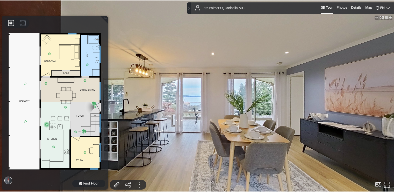 Interior of a modern, bright living and dining area with large windows overlooking a natural landscape, a dining table set with plates and cups, a black sideboard, artwork on the wall, and a kitchen with a breakfast bar.
