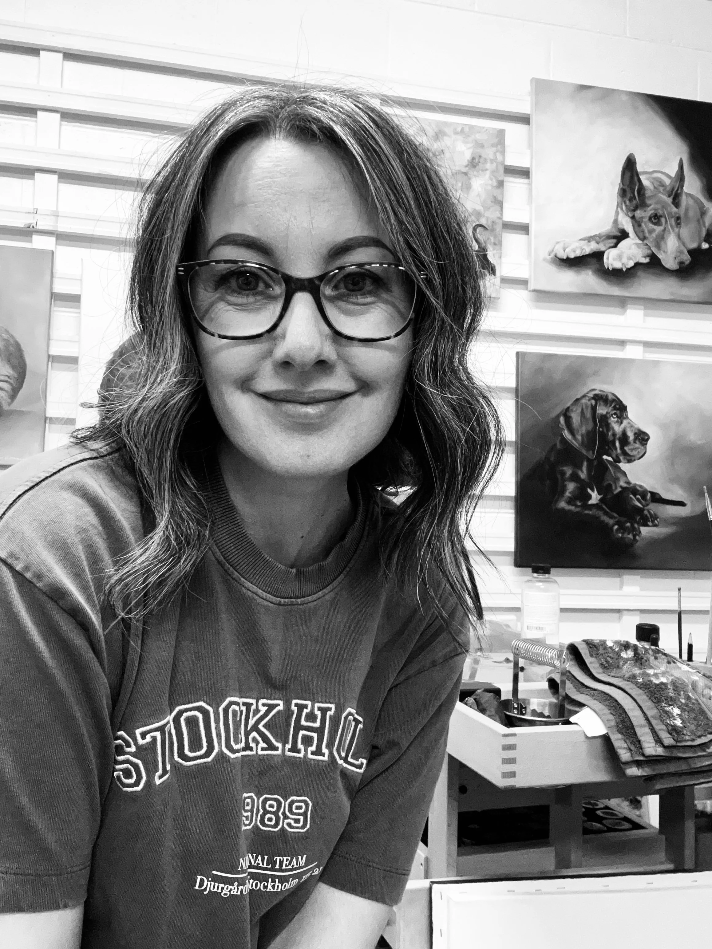 Photo of Jen Sweeney in her art studio, with paintings of dogs on the wall behind her.