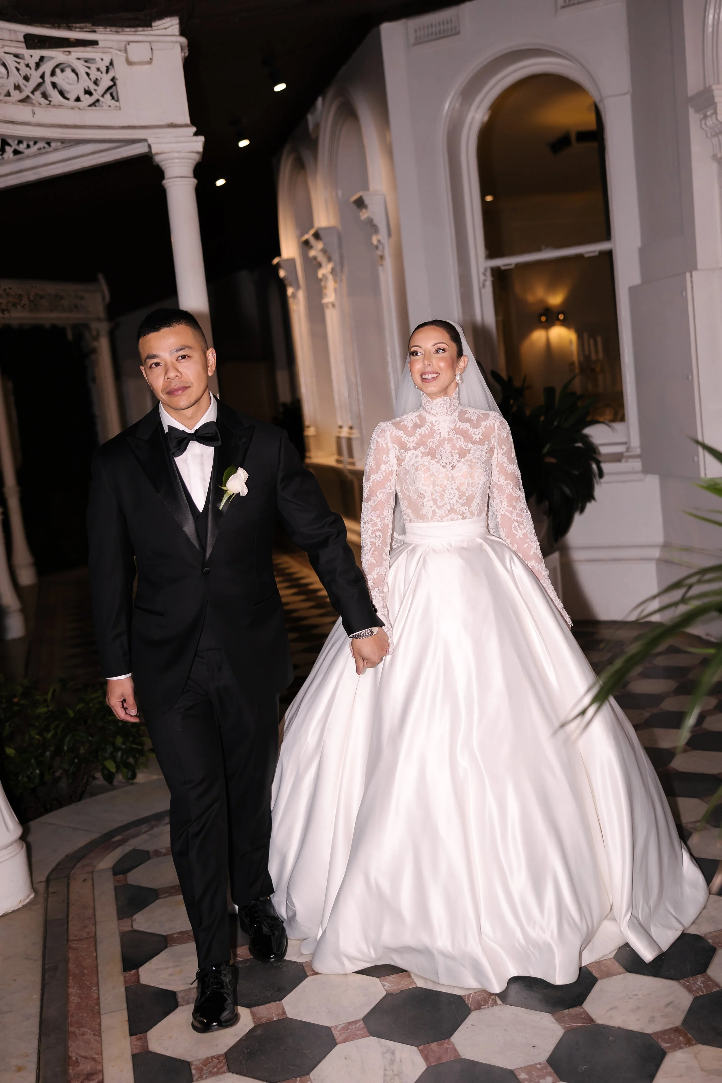 A bride and groom holding hands in wedding attire are walking inside a decorated venue, with the bride wearing a white gown with lace top and the groom in a black tuxedo.
