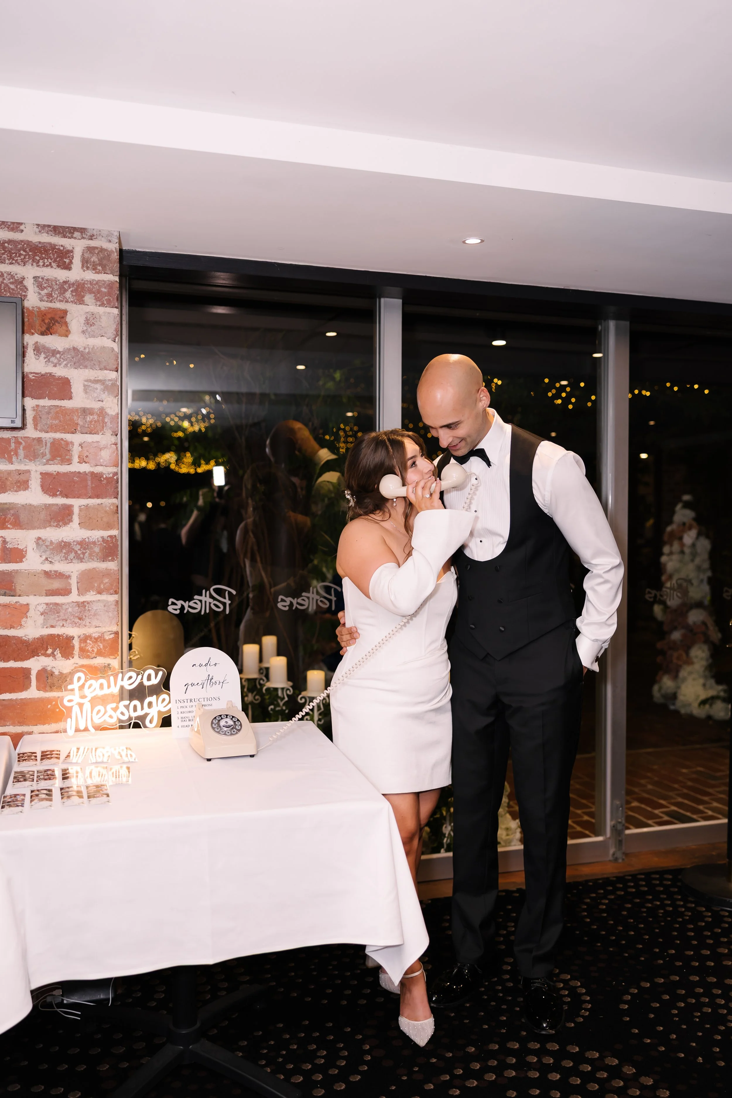 A bride and groom share a playful moment at their wedding reception, with the bride holding a vintage telephone receiver to her ear while the groom smiles beside her, in a decorated venue with candles and string lights outside.
