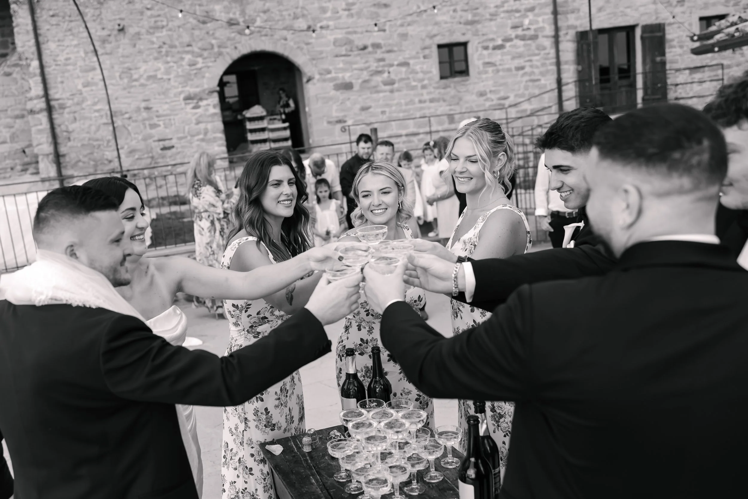 A group of people in formal attire raising glasses for a toast at an outdoor celebration, with a stone building in the background.
