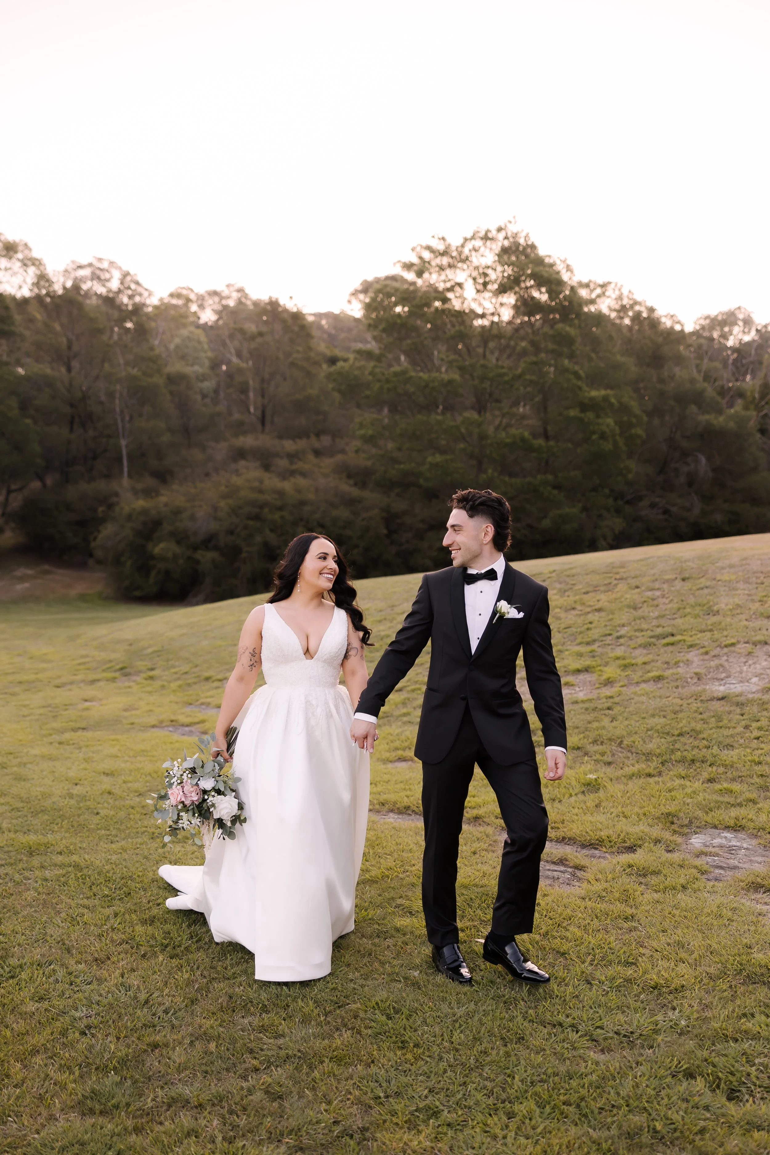 A newlywed couple holding hands and walking on a grassy field during sunset, with trees in the background.