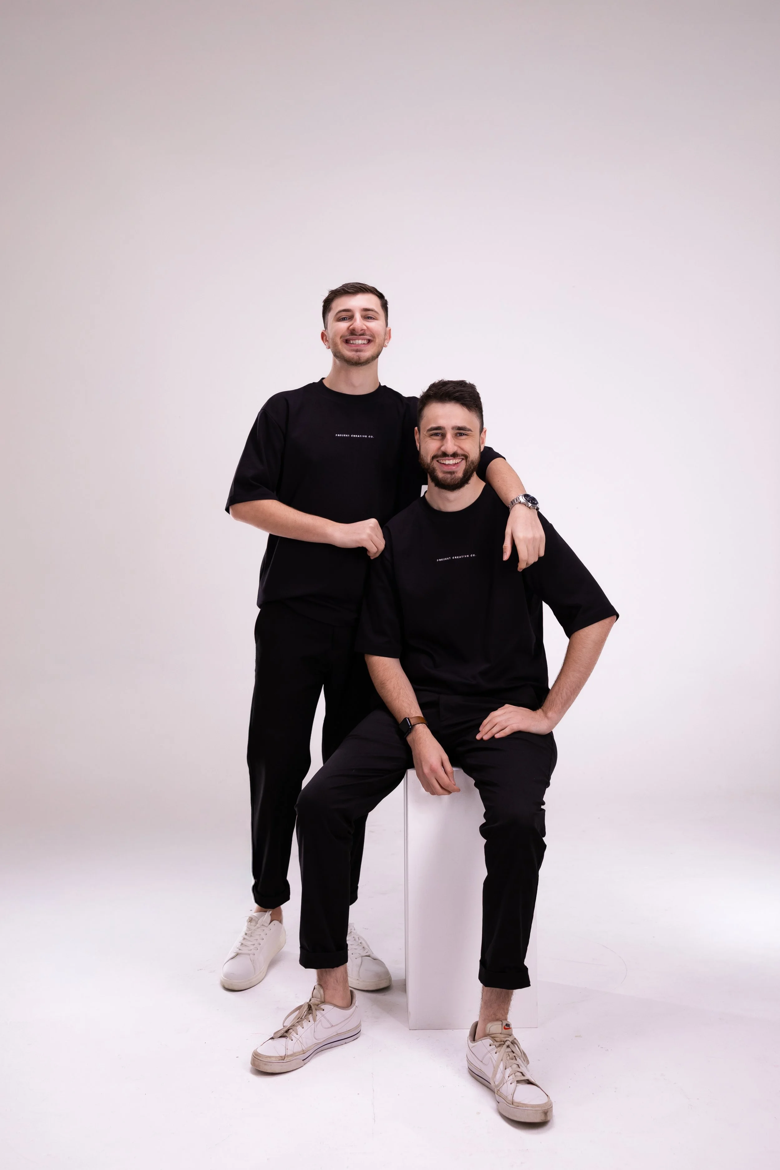Two young men in black t-shirts and pants smiling, one sitting on a white block and the other standing with his arm around the seated man's shoulder, against a plain white background.