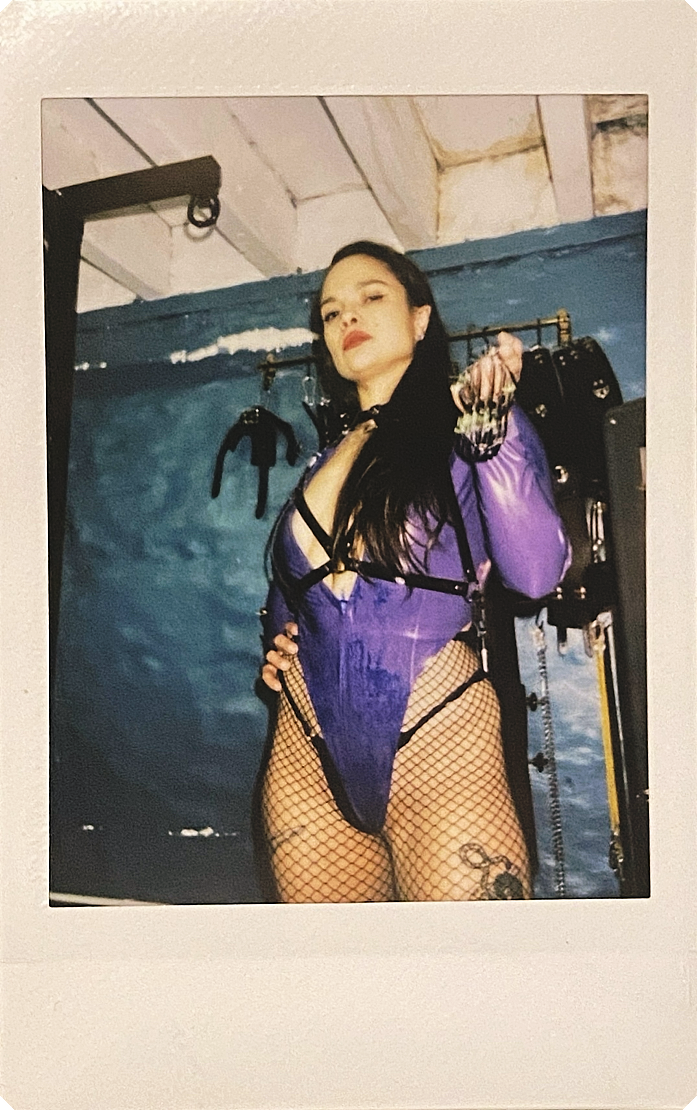 Goddess Jessica dressed in purple lingerie with fishnet stockings, wearing gloves and harnesses, posing confidently in front of a blue wall with a cage.