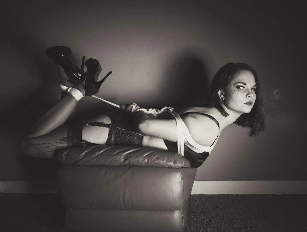 Goddess Jessica with dark hair and makeup lying on her stomach on a couch, wearing high heels, fishnet stockings, and lingerie, with her hands tied behind her back and a rope around her waist, against a plain wall. Dominate stare. 
