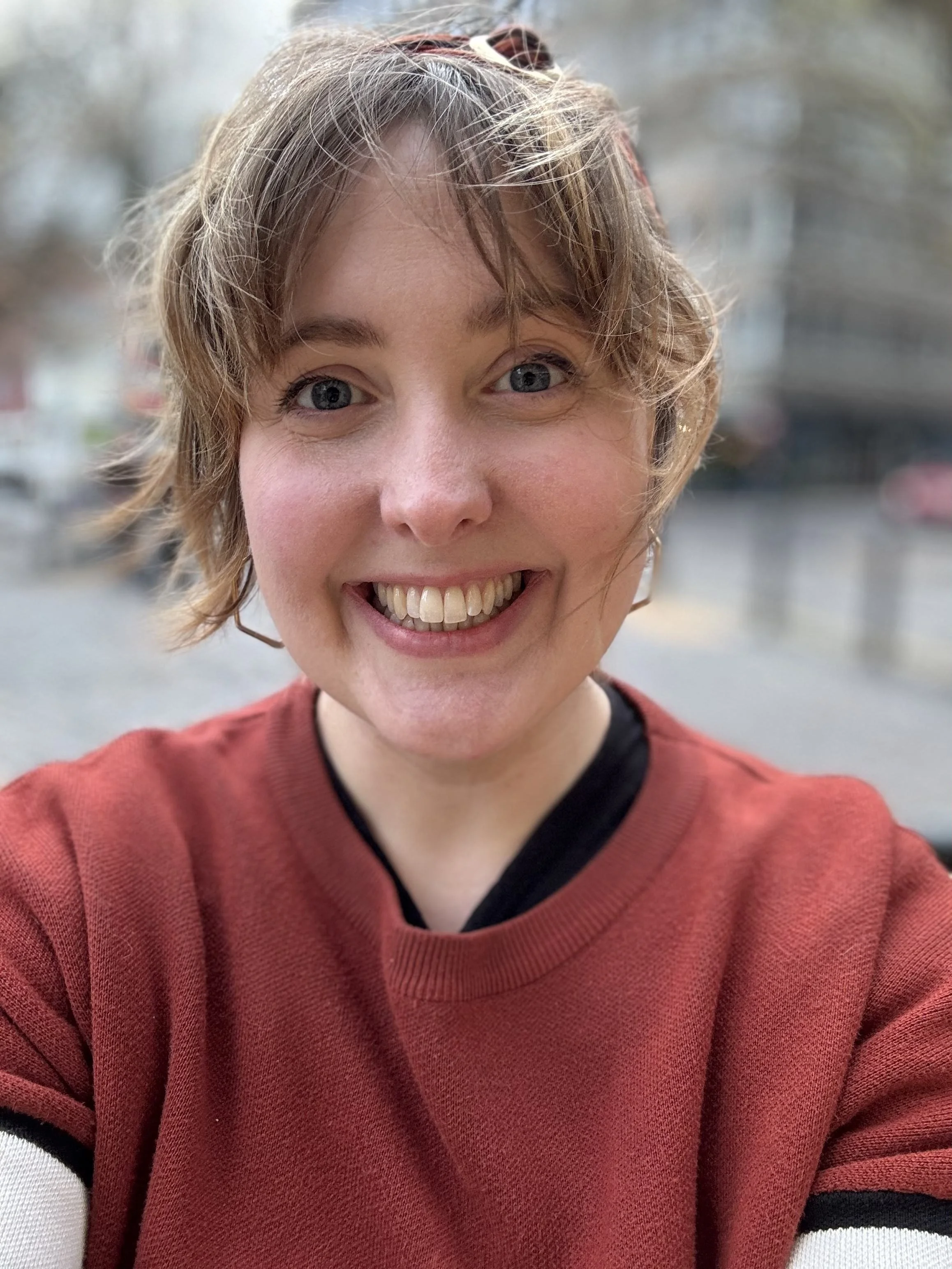 A woman with a messy fringe blowing in the wind, smiling outdoors during daytime, wearing a red sweater.