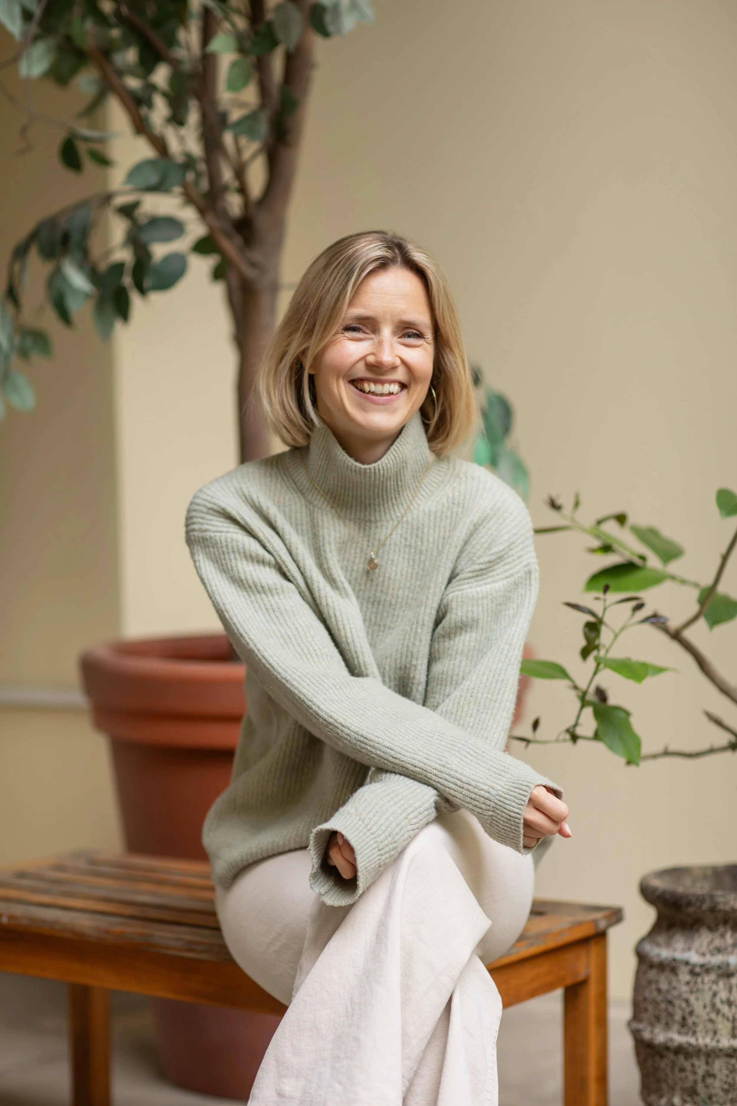 A smiling woman with blonde hair sitting on a wooden bench in front of a large potted plant with green leaves, wearing a beige sweater and light-colored pants.