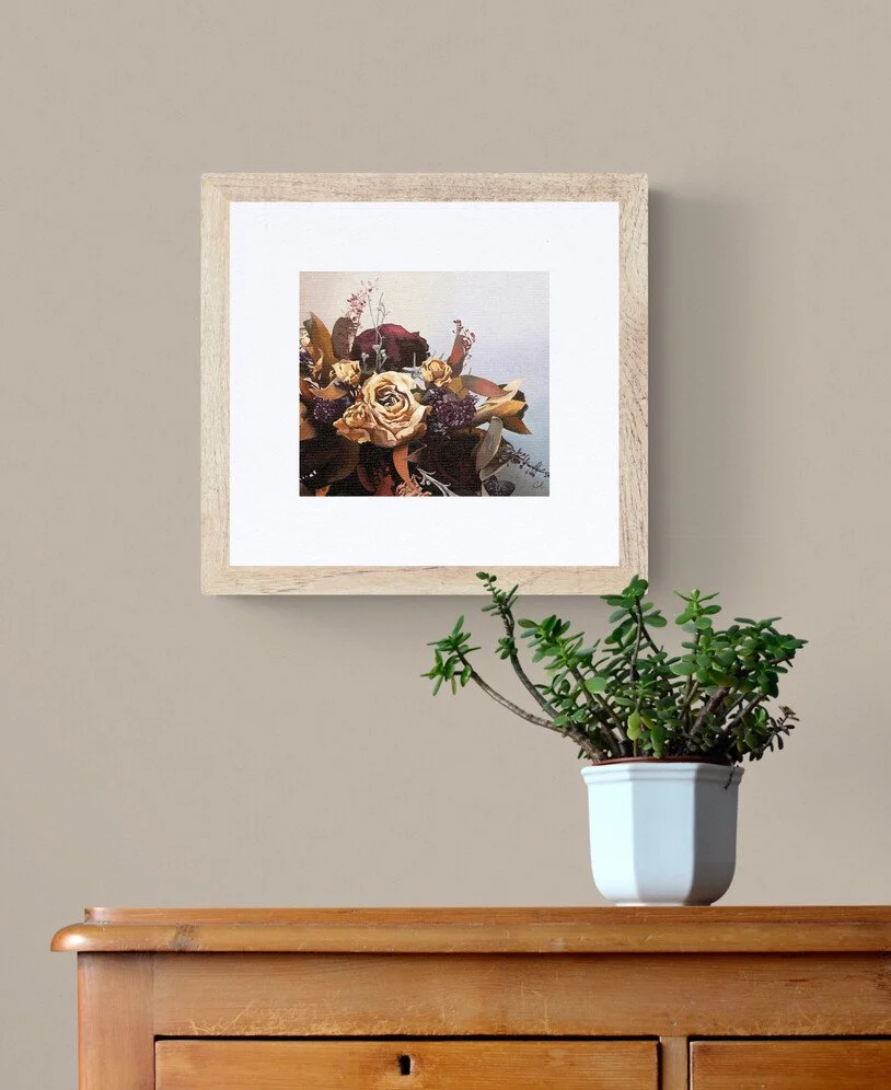 A framed painting of a floral bouquet hangs on a beige wall above a wooden surface, with a potted succulent plant in a white pot placed on the surface.