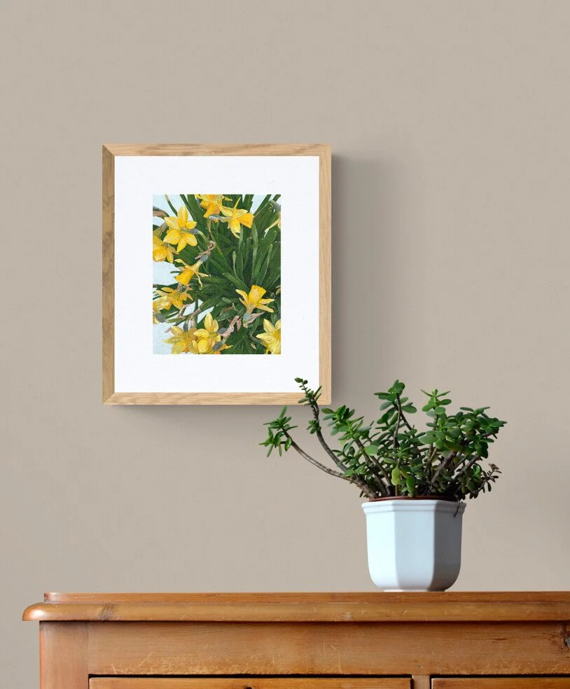 A potted succulent plant with green, paddle-shaped leaves sitting on a wooden surface. Behind it, a framed picture of yellow flowers with green leaves hangs on a beige wall.