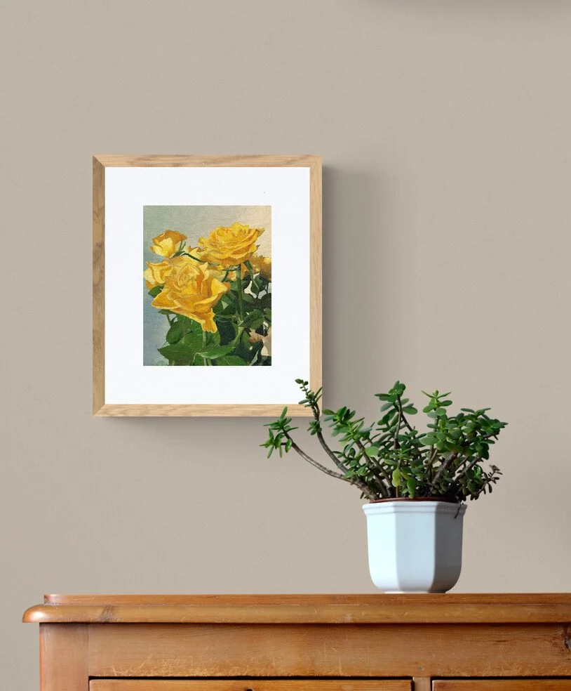 A framed painting of yellow roses hangs on a beige wall, with a potted jade plant on a wooden surface below.