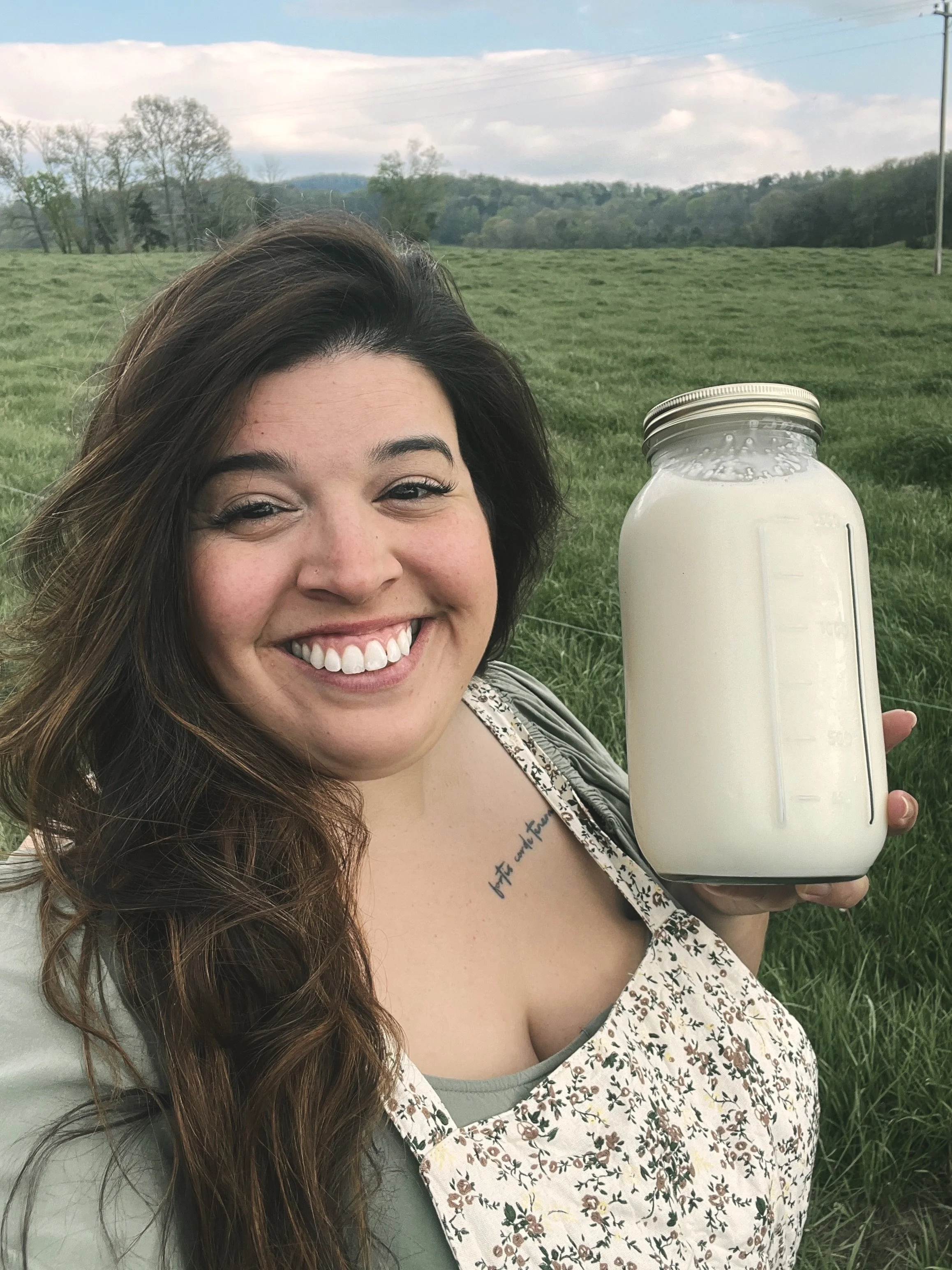 A woman with long brown hair smiling and holding a large glass jar with milk in a green field with trees and hills in the background.