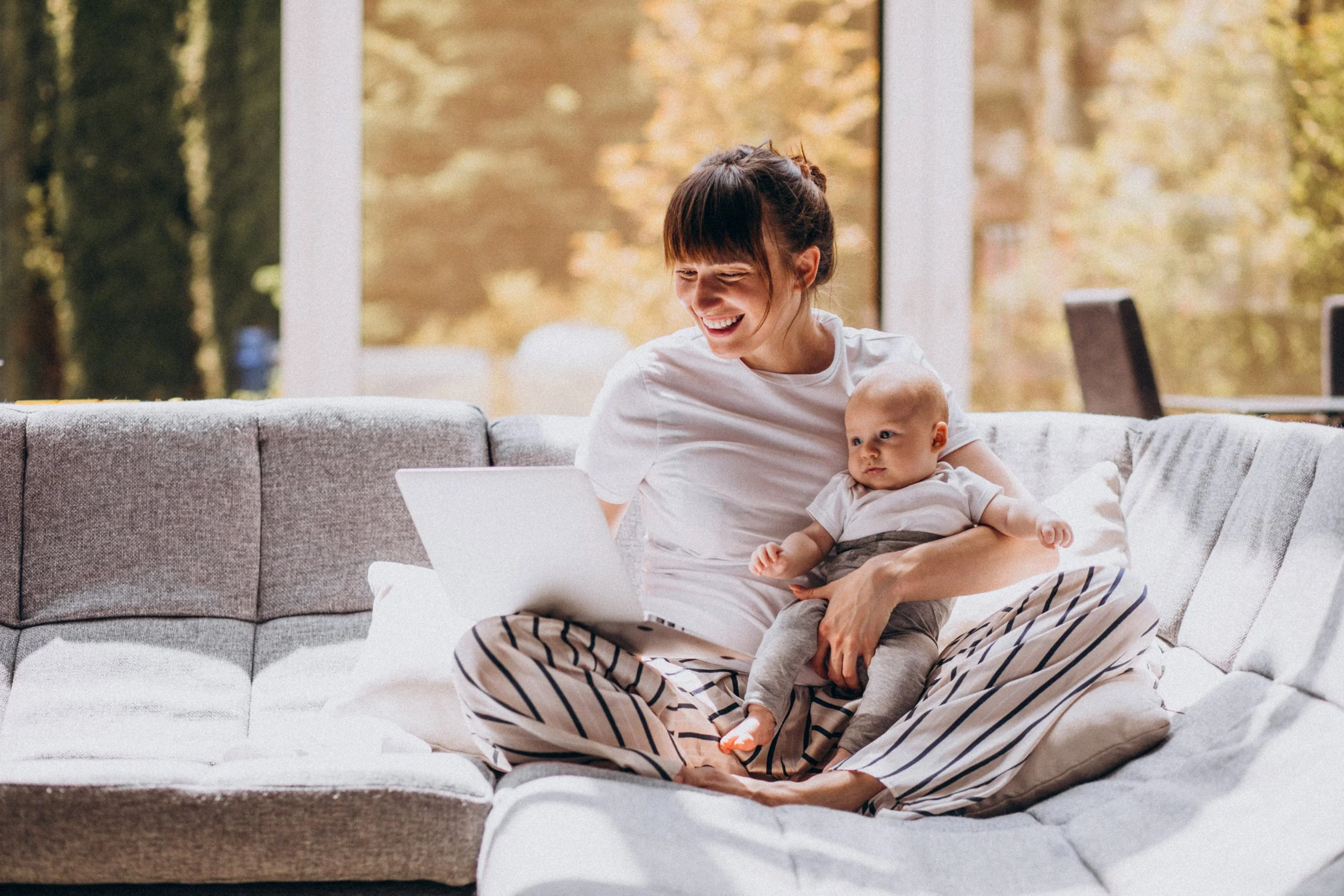 A woman sitting on a couch with a baby, using a laptop, in a sunlit room with large windows and autumn trees visible outside.