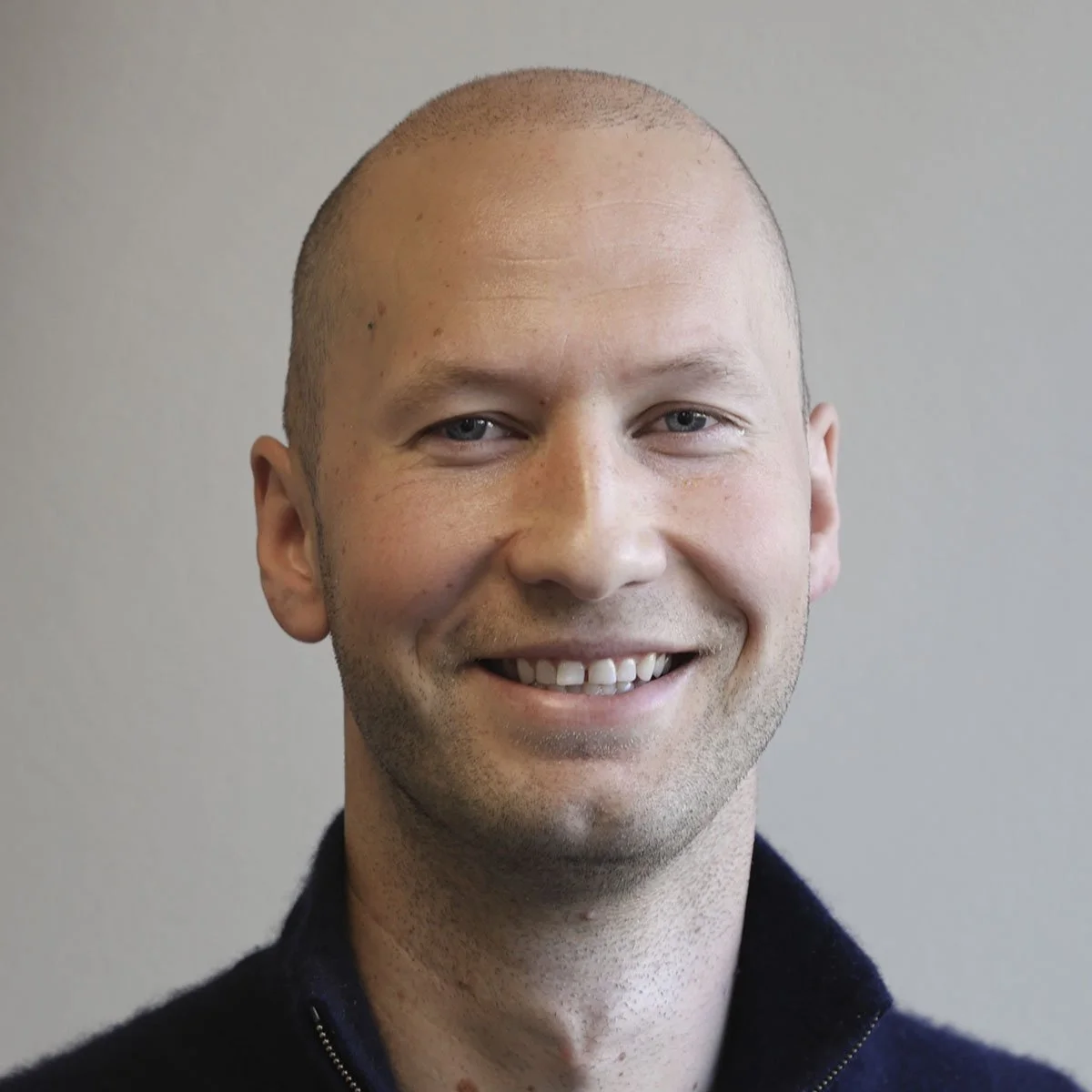 Un mann med kort hår og smil, kledd i svart genser, poserer foran en grå vegg.