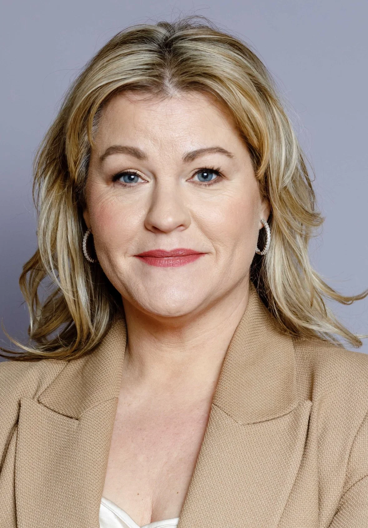Portrait of a Jo Tanner with blonde hair, wearing a tan blazer and earrings, against a gray background.