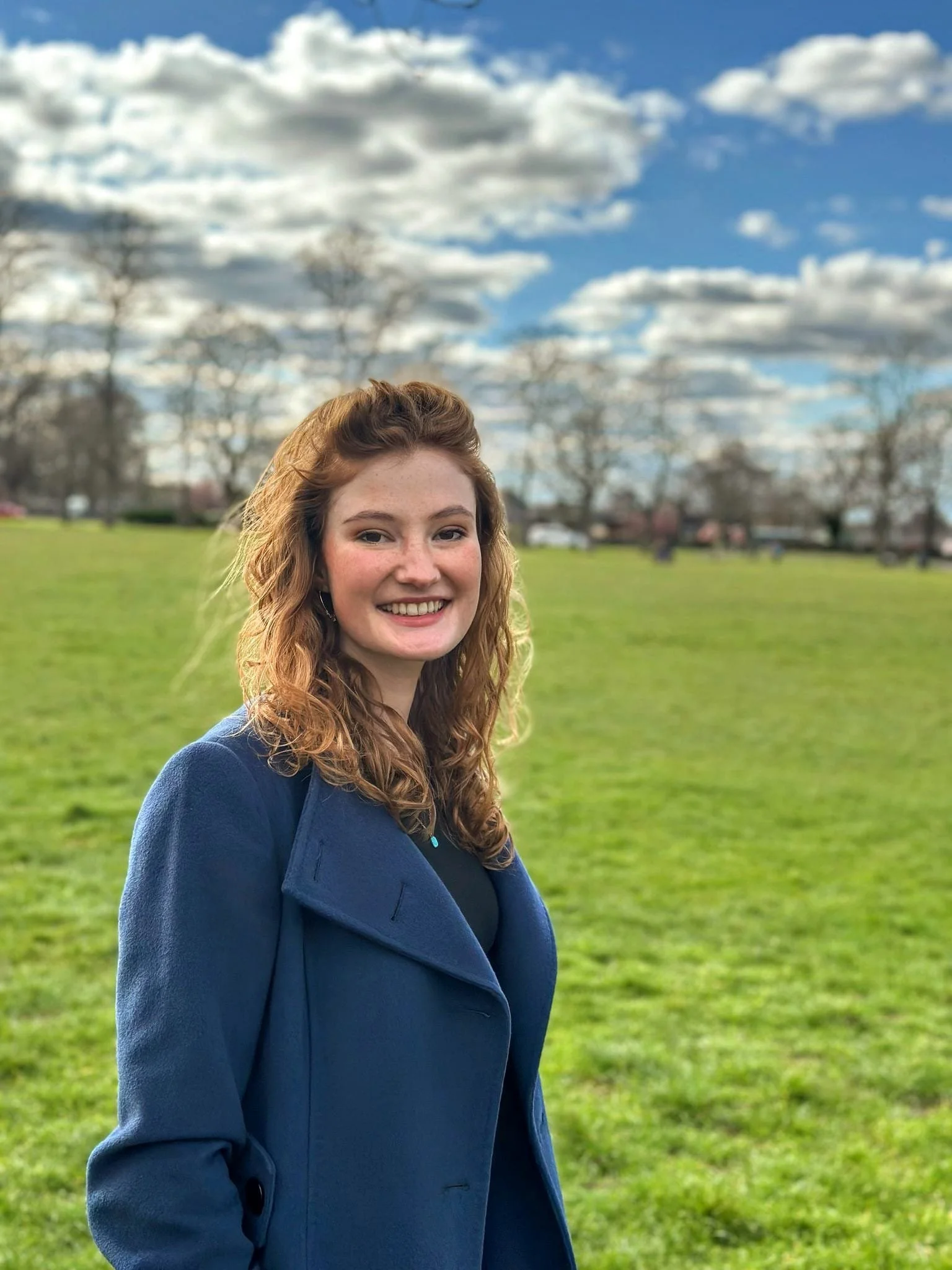 A young woman with curly red hair, smiling, wearing a blue coat, standing outdoors on a grassy field with trees and a partly cloudy sky in the background.