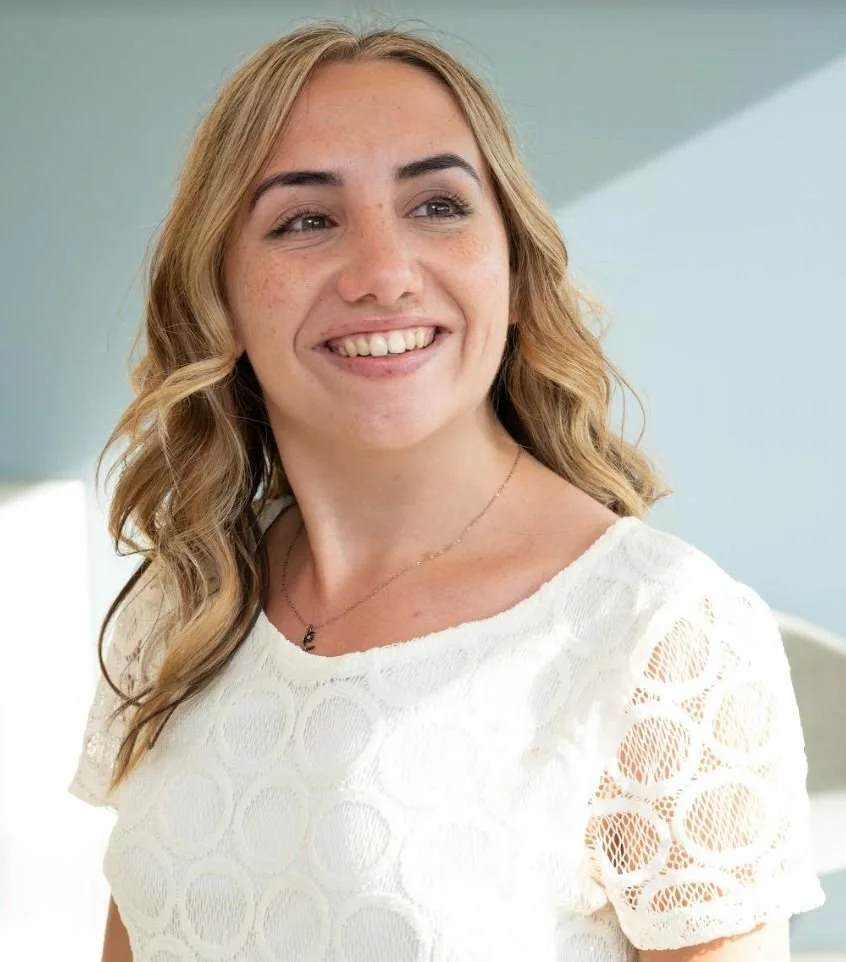 A young woman with wavy blonde hair smiling and looking to the side, wearing a white textured top and a delicate necklace.