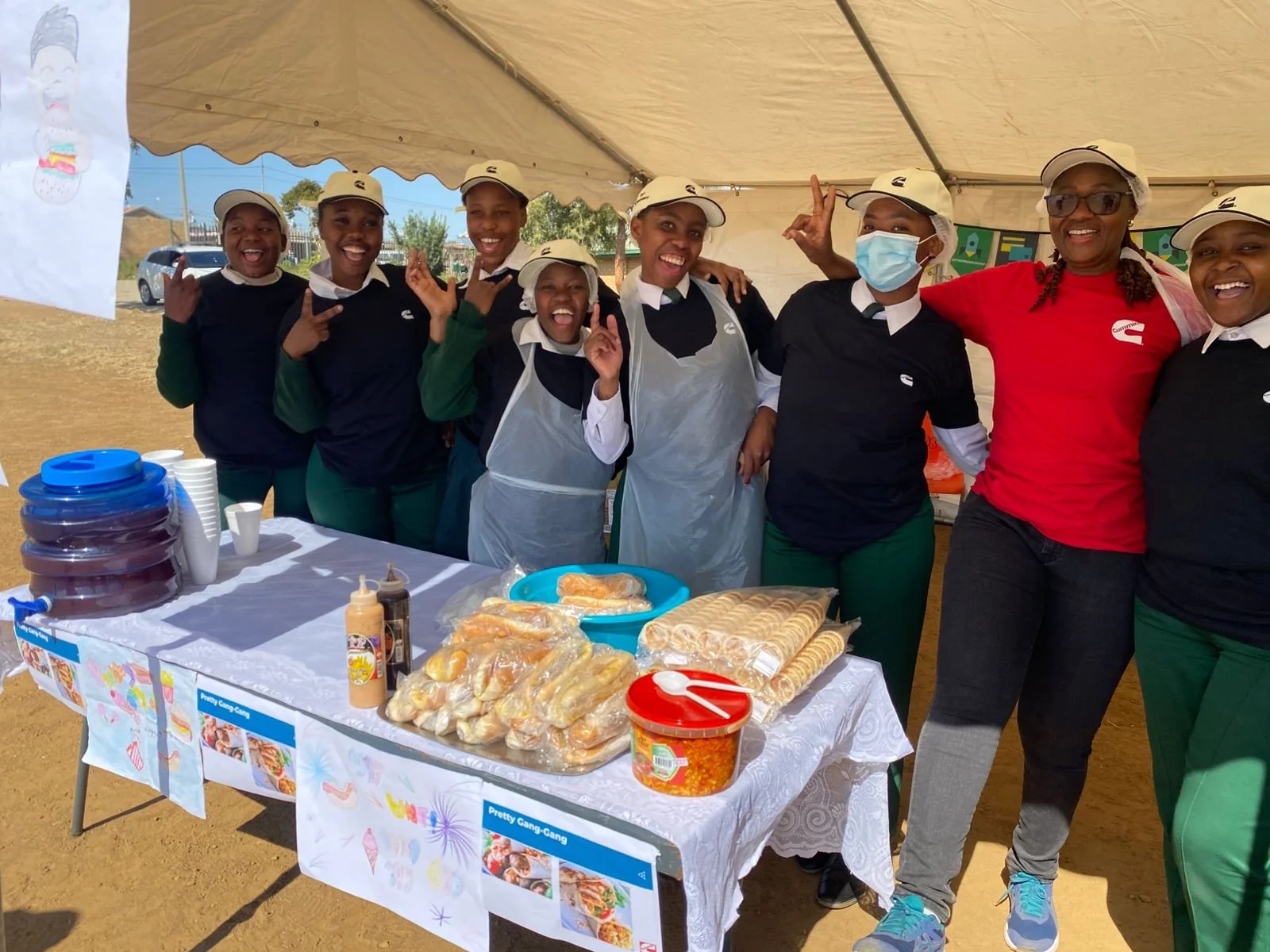 Group of women standing behind a food table at an outdoor event, smiling and posing for the photo.