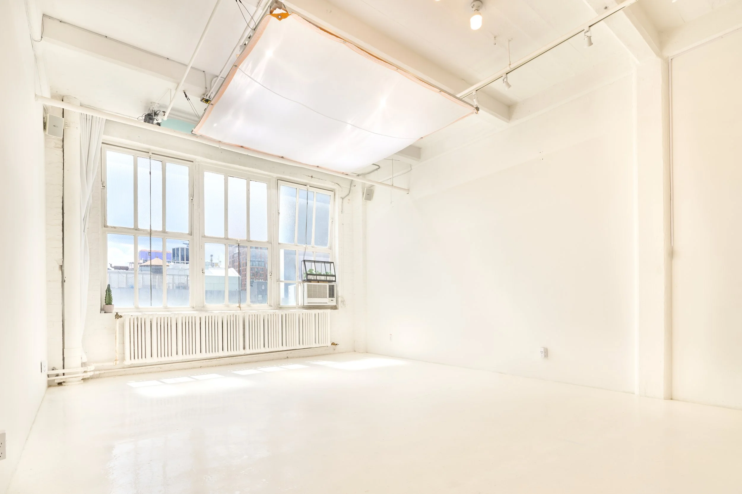 An empty room with white walls, a large window with multiple panes, a radiator beneath the window, and an air conditioning unit. There is a small cactus plant on the windowsill. The ceiling has a large patch of exposed drywall with lighting fixtures.