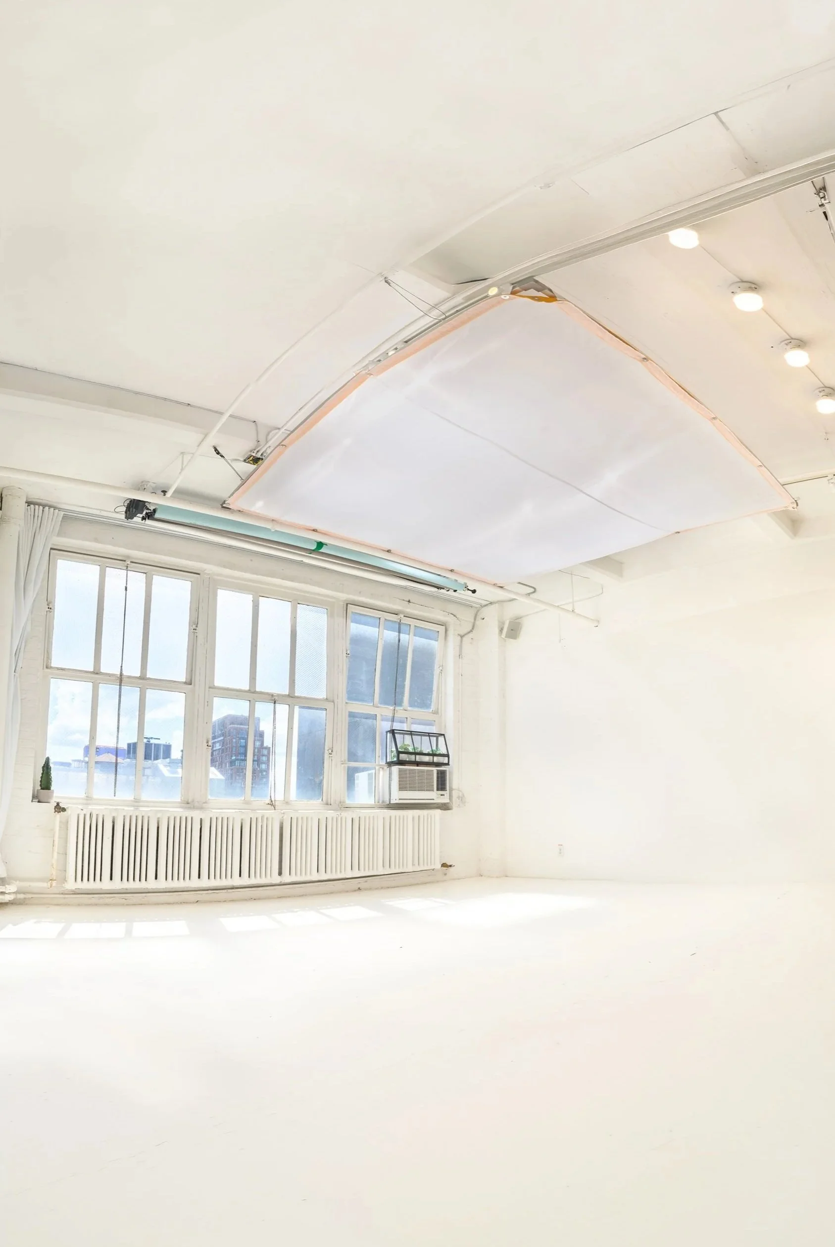 A bright studio with large windows, a white radiator below, and an air conditioning unit. The ceiling features a large, suspended, rectangular panel with lighting fixtures. Outside city buildings are visible through the windows.