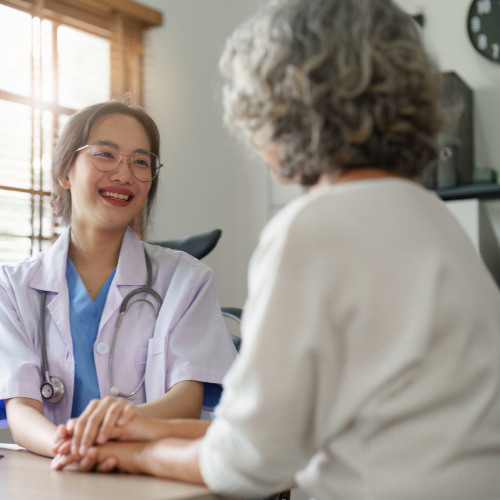 A young female healthcare professional is smiling and holding hands with an elderly woman in a medical office.