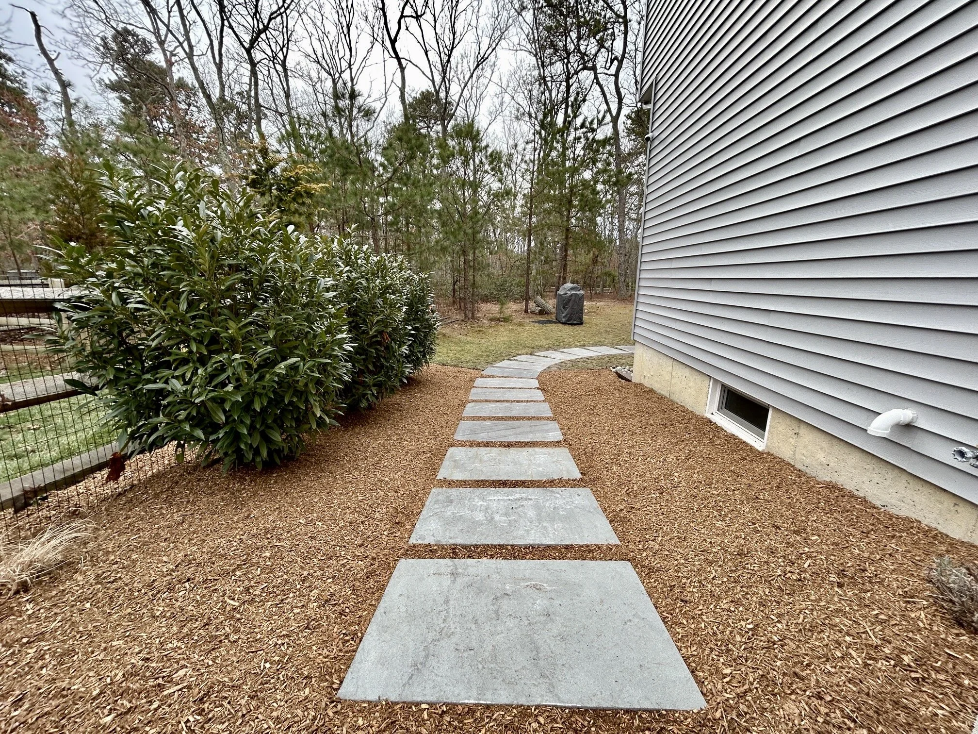 Concrete stepping stones forming a curved pathway beside a house with gray siding, mulch ground cover, and a green bush on the left, with a wooded area in the background.
