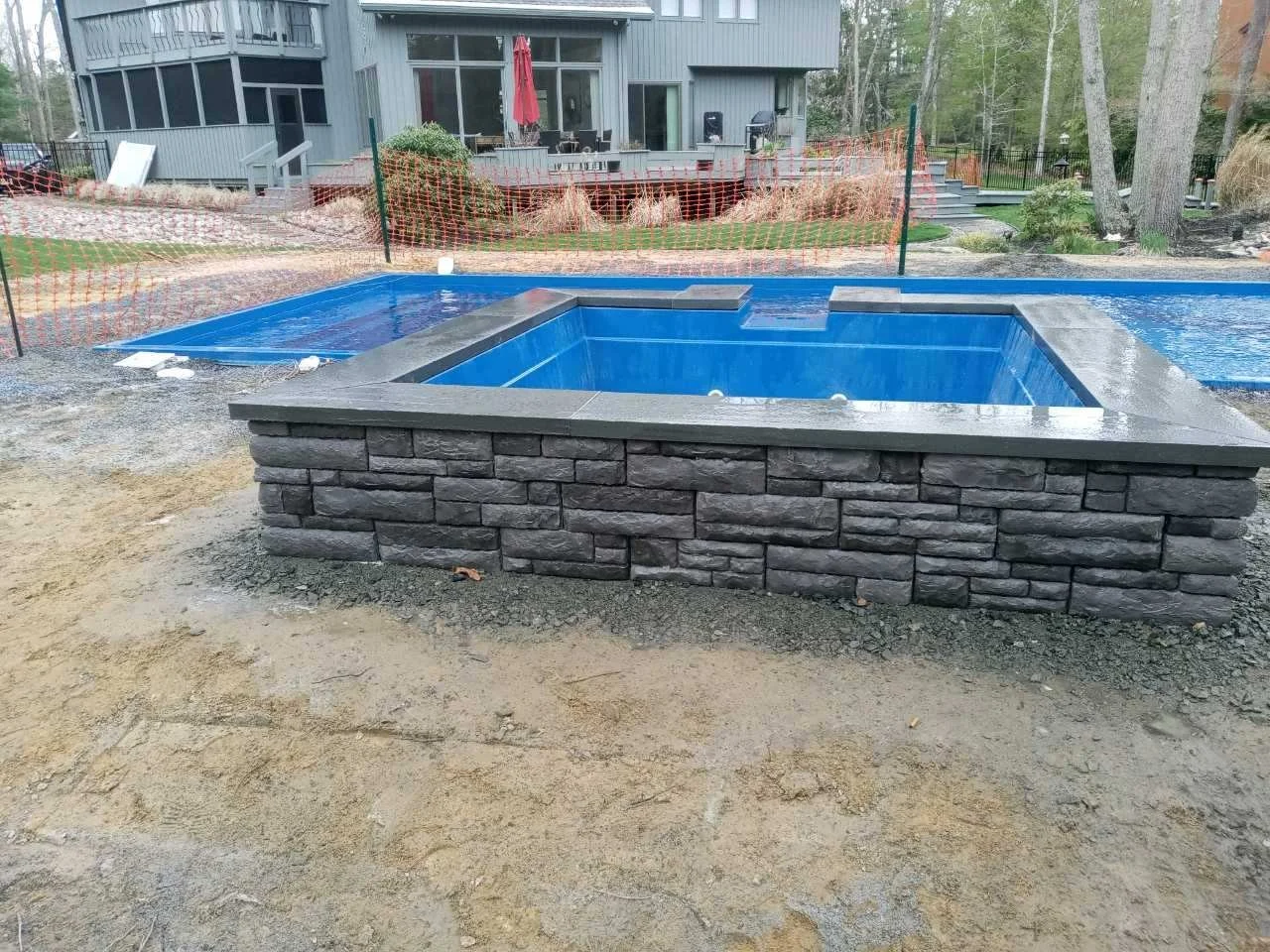 In-ground rectangular pool with blue interior, surrounded by a stone deck, in a backyard with construction barriers and a house in the background.