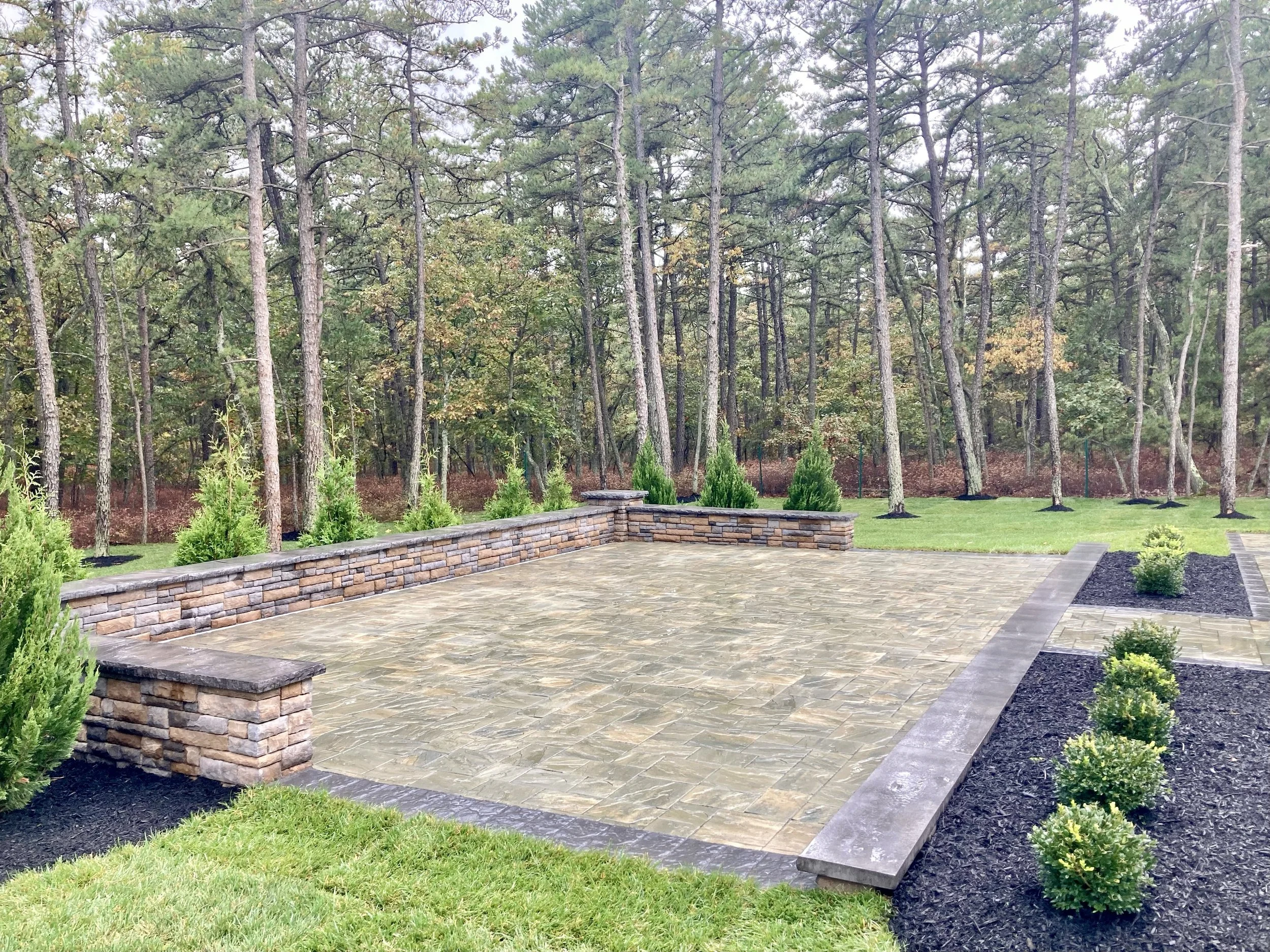 A landscaped backyard with a paved area, low brick wall, small shrubs, and a forest background with tall trees.