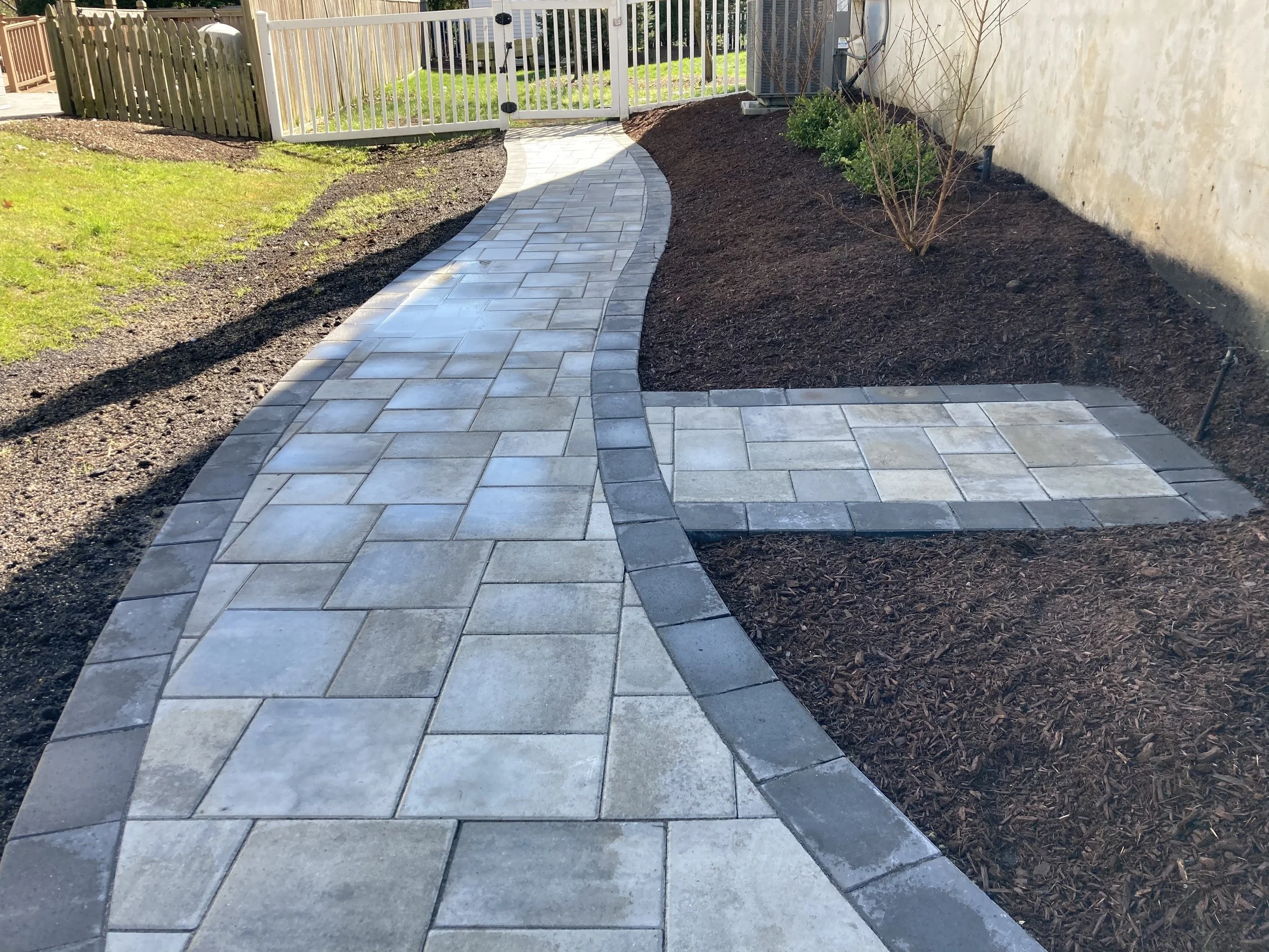 A backyard pathway with gray and tan rectangular pavers, curved in design, leading to a white gate at the end. Surrounding the pathway are freshly mulched garden beds with small plants and an area of green grass.