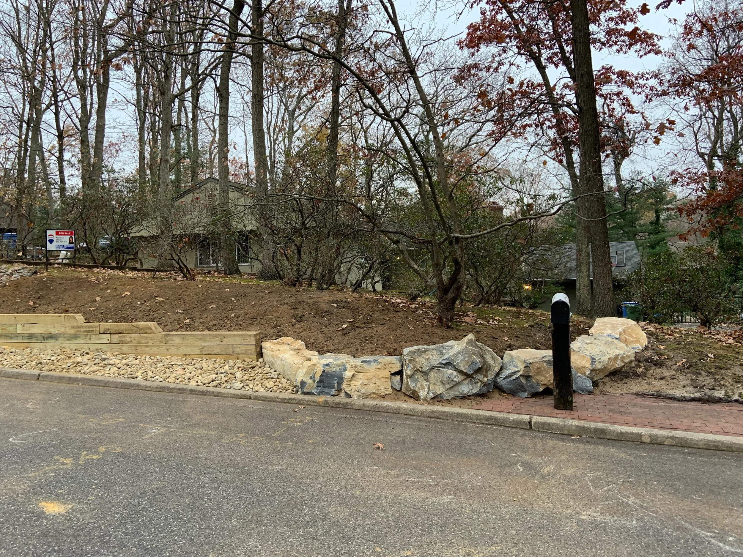 A roadside scene with a black mailbox in front of a landscaped area with large rocks and new wooden retaining wall, surrounded by leafless trees and residential houses in the background on an overcast day.