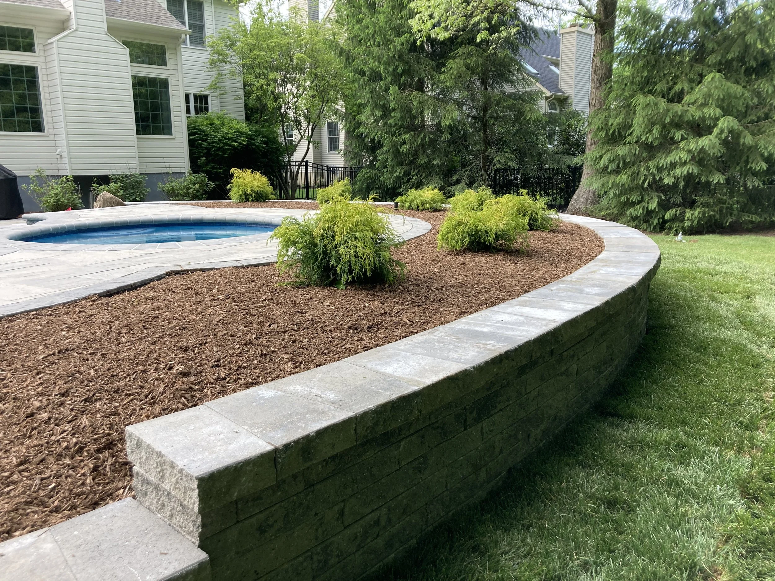 A backyard garden with a curved stone retaining wall, mulch, small green shrubs, a swimming pool, and surrounding trees and houses.