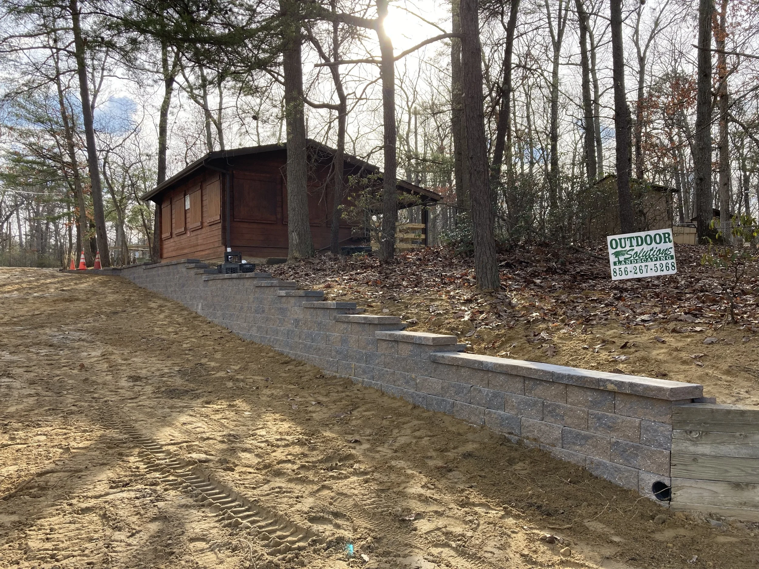 A newly built brick retaining wall on a sandy slope in front of a brown wooden house surrounded by trees in a wooded area, with a sign for outdoor landscaping services.