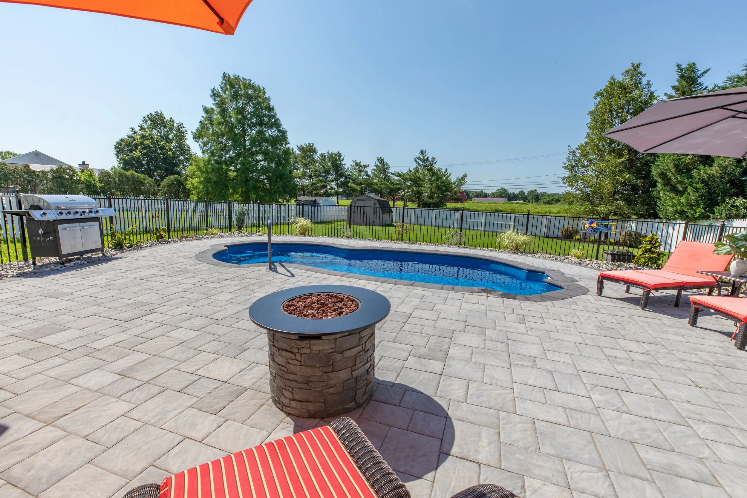 Backyard with a swimming pool, lounge chairs with red cushions, patio umbrellas, a fire pit, and a barbecue grill, surrounded by a black fence and green trees under a clear blue sky.
