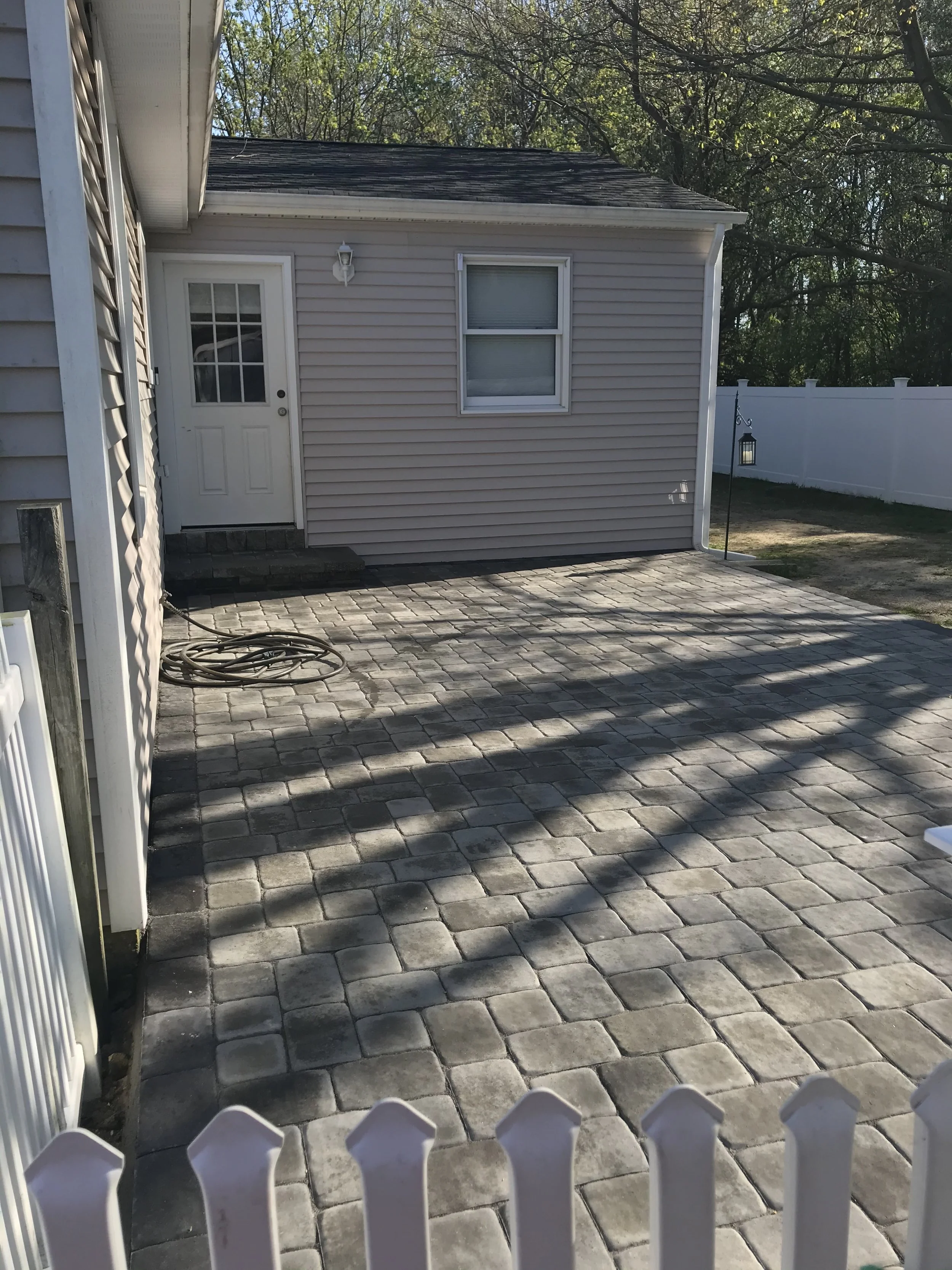 Backyard patio with interlocking stone pavers, house with off-white siding, white door and window, white fence, hanging lantern, garden hose on the ground, surrounded by trees.