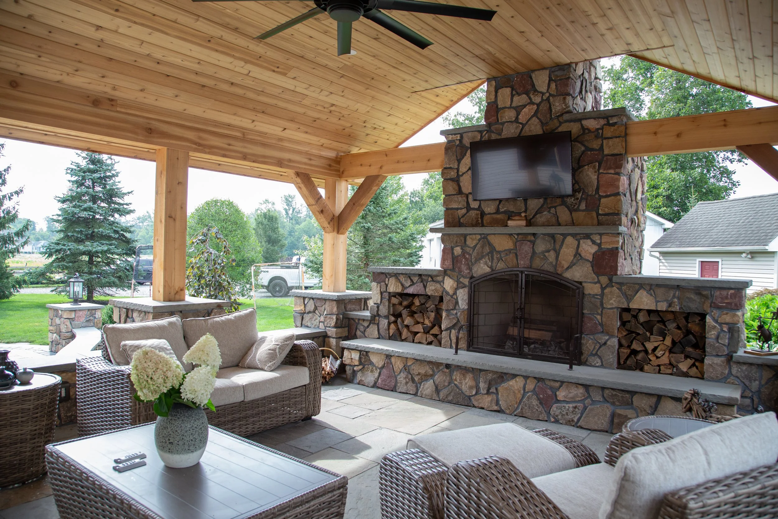 Covered outdoor patio with wicker furniture, a stone fireplace with a TV mounted above, wood storage on either side, and a ceiling fan, overlooking a lush backyard with trees and a house.