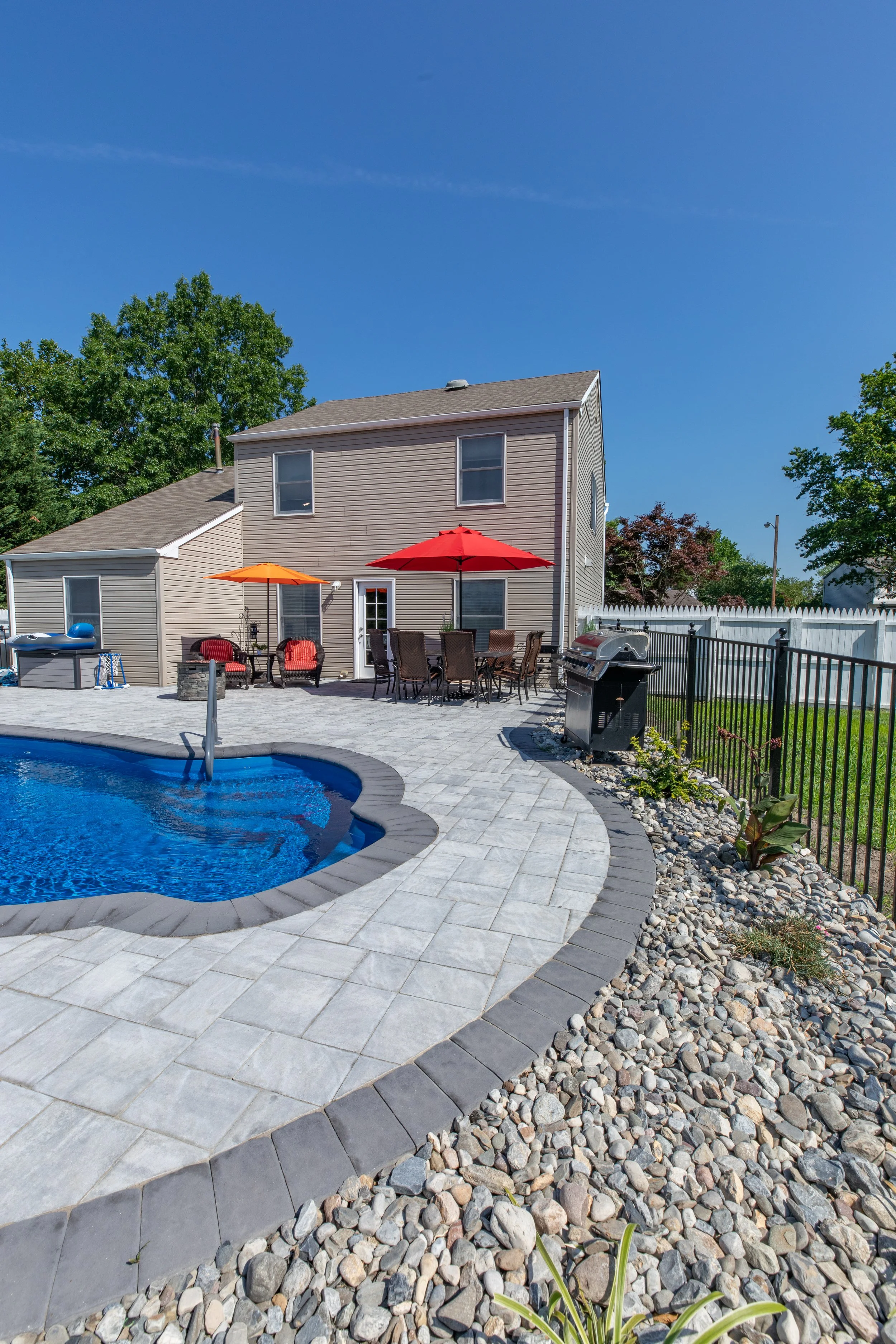 Backyard with in-ground swimming pool, outdoor furniture with umbrellas, grill, and a house with beige siding, surrounded by a white fence and green trees.
