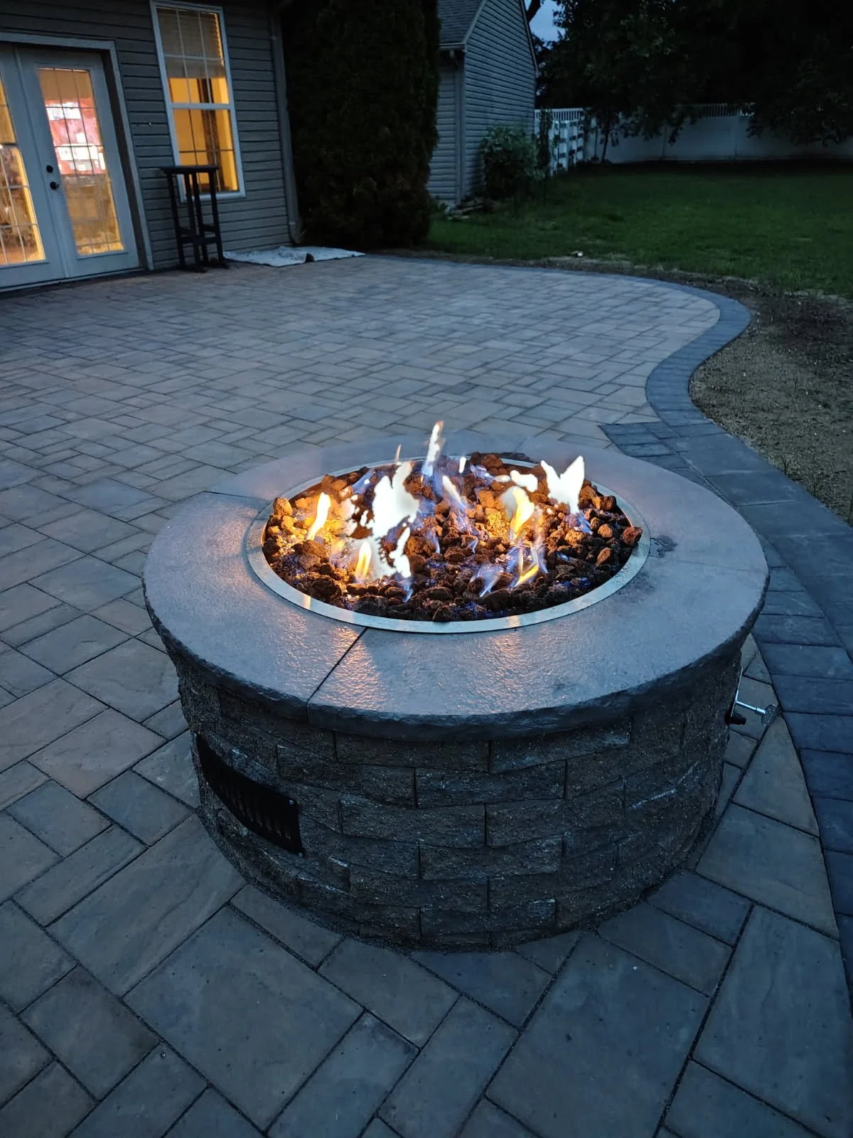 A fire pit with flames burns on a gray stone base in a backyard during dusk, with paver stone flooring and a house with glass doors and windows in the background.