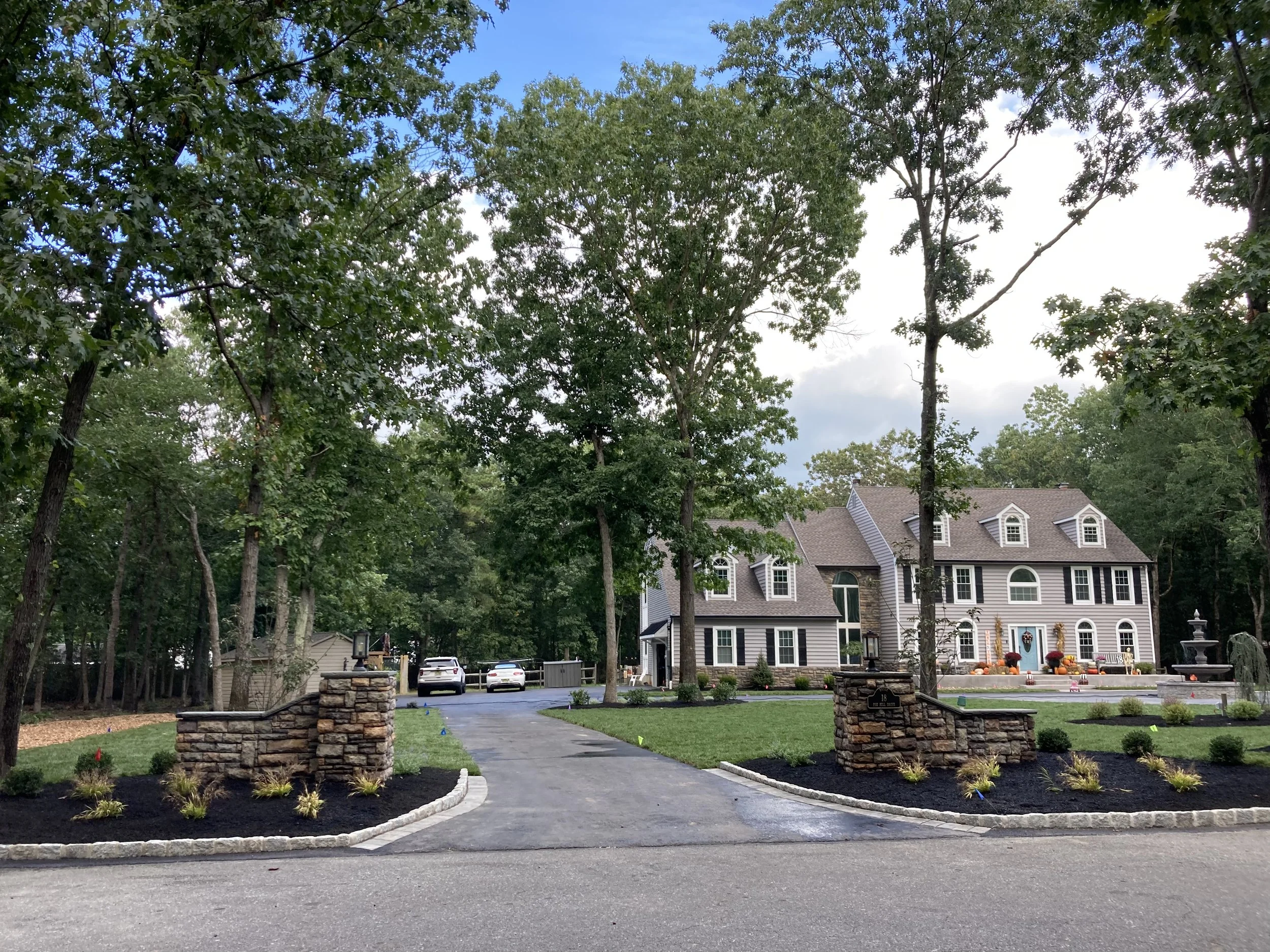 A large, modern house with light gray siding and multiple gabled roofs, surrounded by a landscaped yard with a driveway and trees.