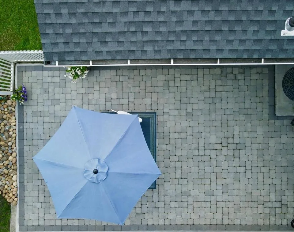 Overhead view of a backyard patio area with a large blue patio umbrella, a doormat, a section of a house with gray shingles, a small patch of grass, and decorative rocks.