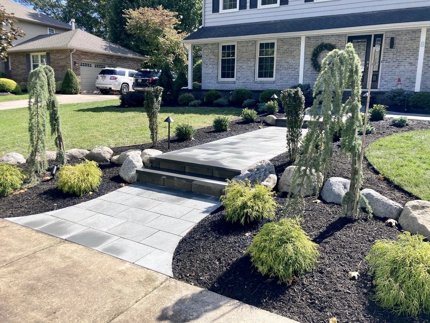 A landscaped front yard of a house with a paved walkway, small bushes, rocks, and young trees, leading to the house front door.