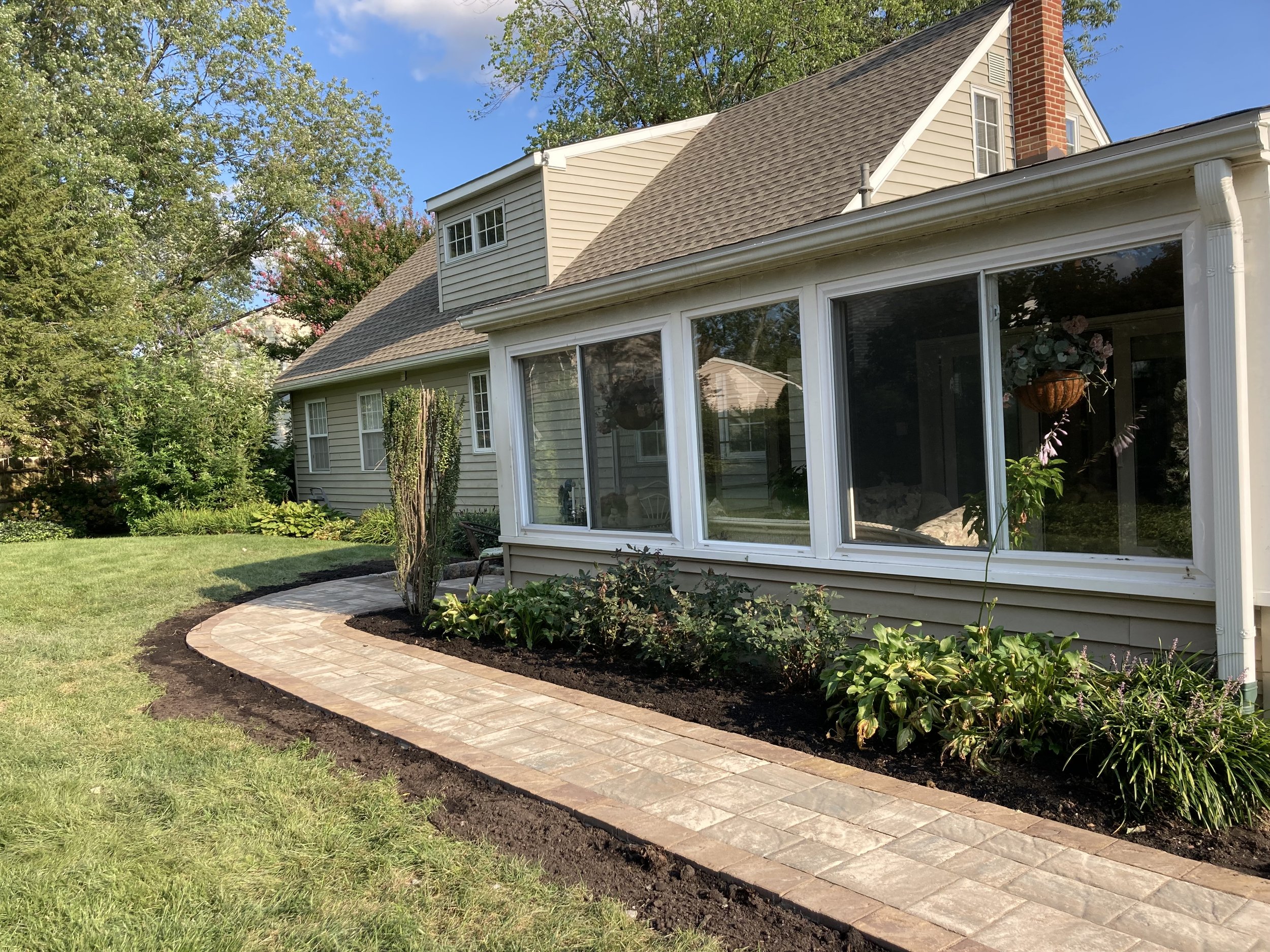 A backyard with a curved brick pathway, green lawn, and landscaped flower bed in front of a house with large sunroom windows. There are trees and a blue sky in the background.