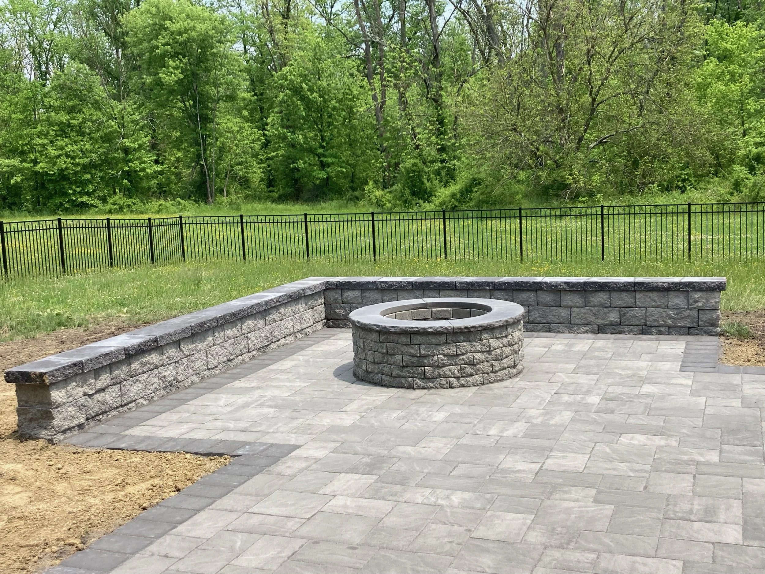 A paved outdoor area with a circular stone fire pit and a stone retaining wall, surrounded by a grassy field and trees, with a black metal fence in the background.