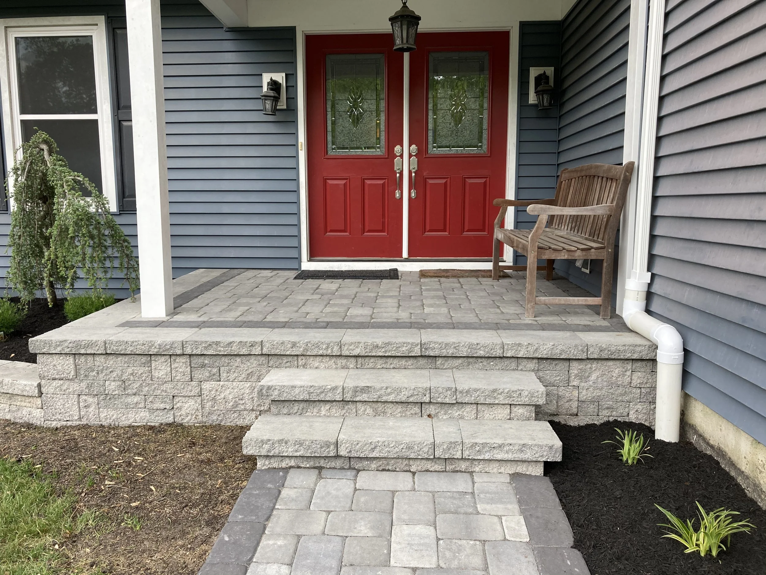 Front porch with red double doors, gray siding, a wooden bench, lantern-style light fixtures, and a small garden with young plants.