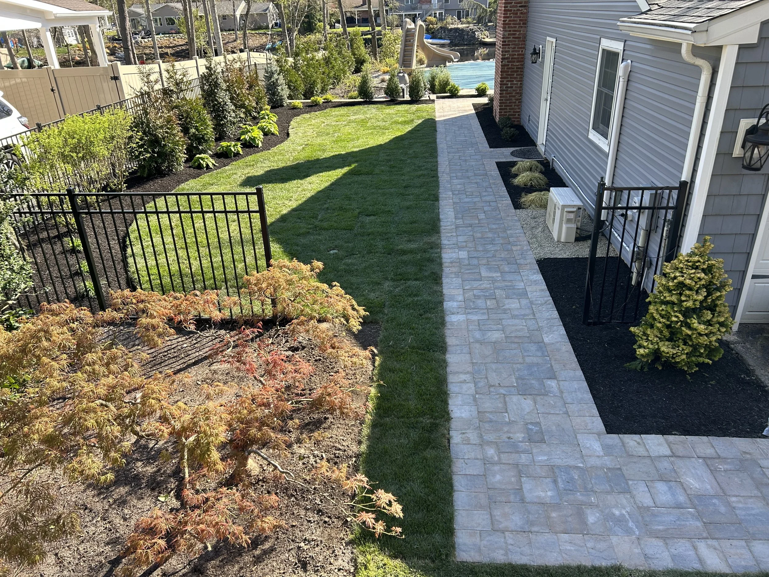 Backyard with a paved walkway, green lawn, shrubs, and a small fence.