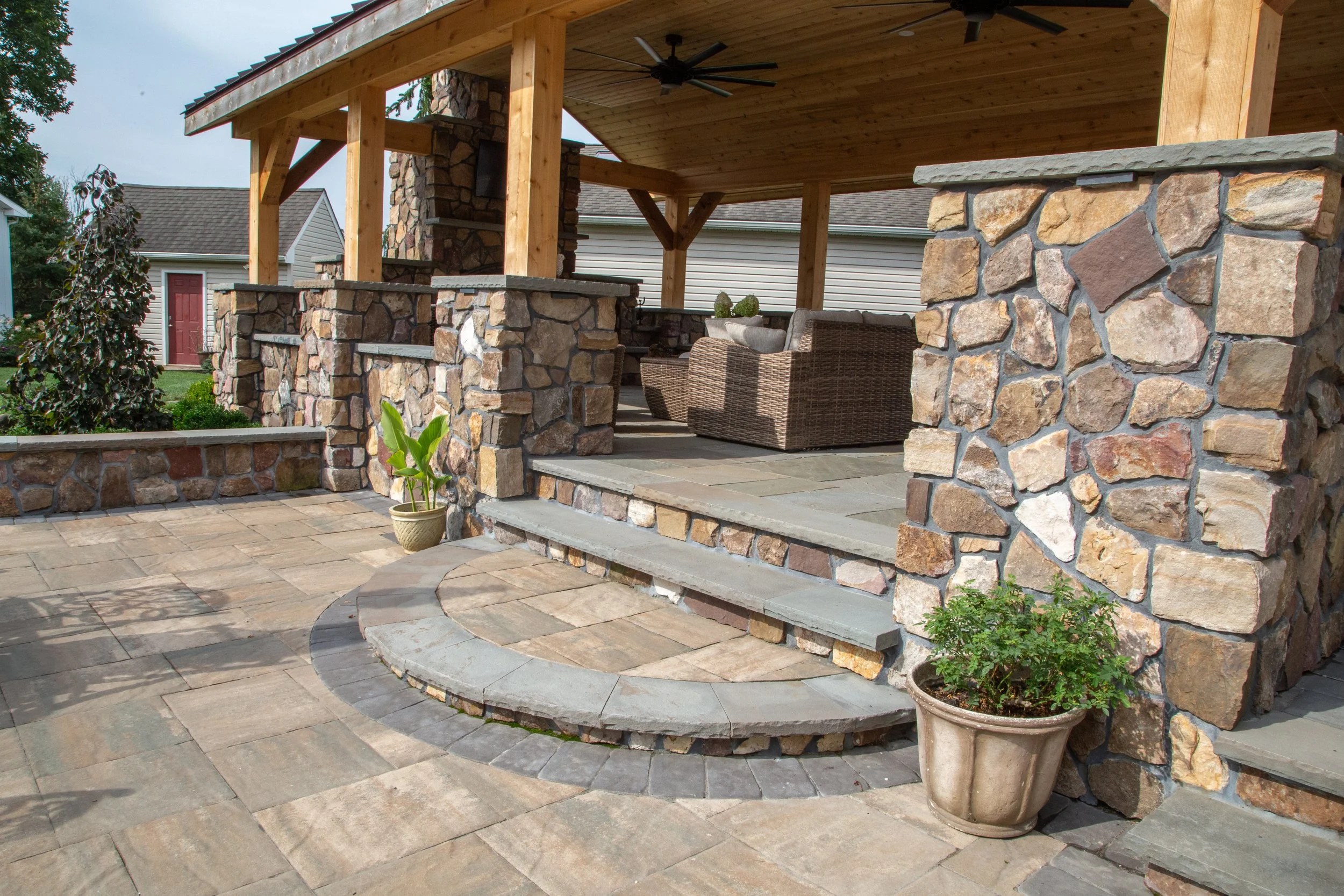 Exterior view of a covered patio with stone and wood construction, featuring steps leading to an outdoor seating area with wicker furniture and decorative potted plants on a paved stone patio.
