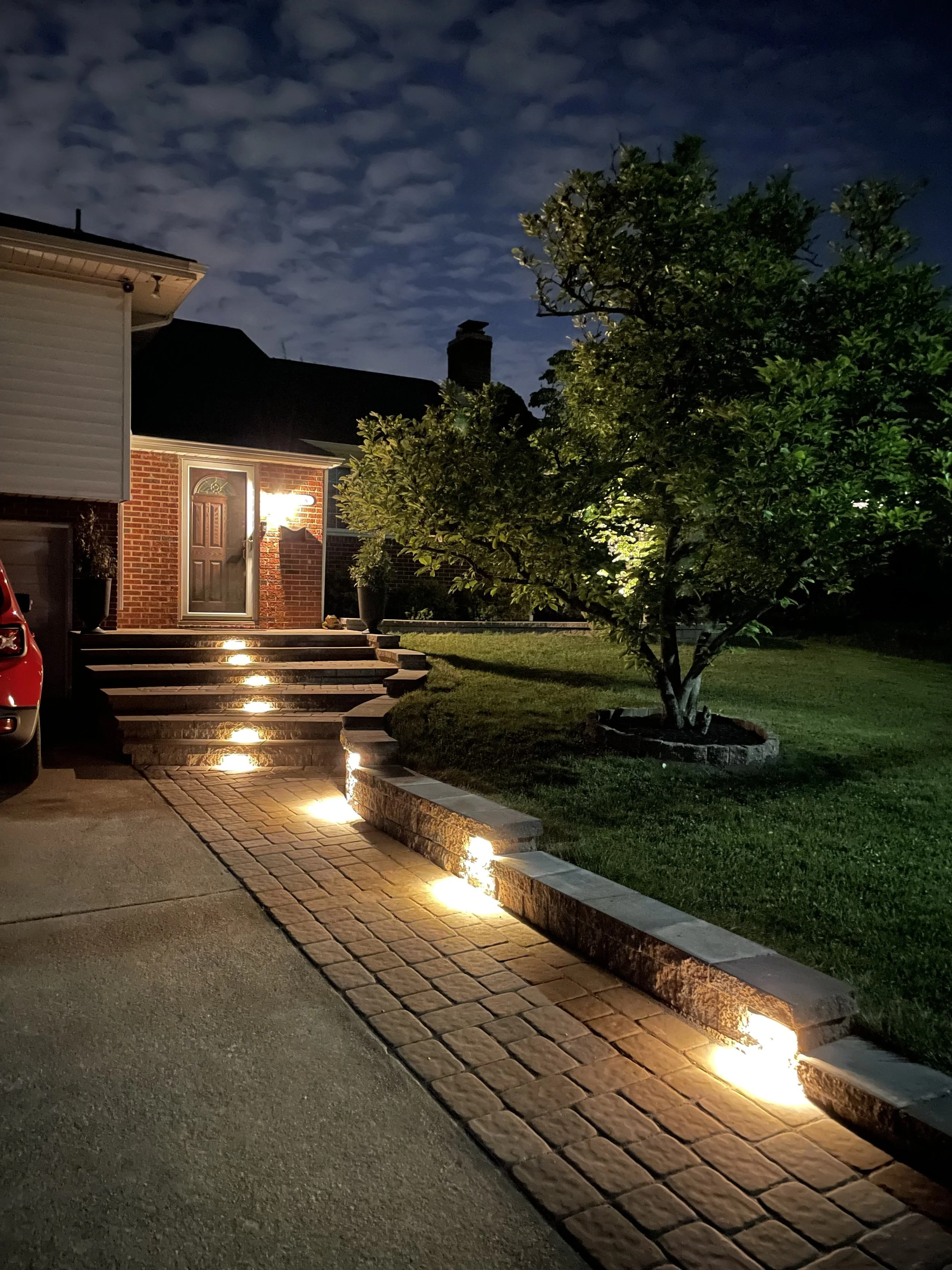 Nighttime view of a house entrance with illuminated pathway lights along the brick stairs and walkway, a green tree on the front yard, and a dark sky with scattered clouds.