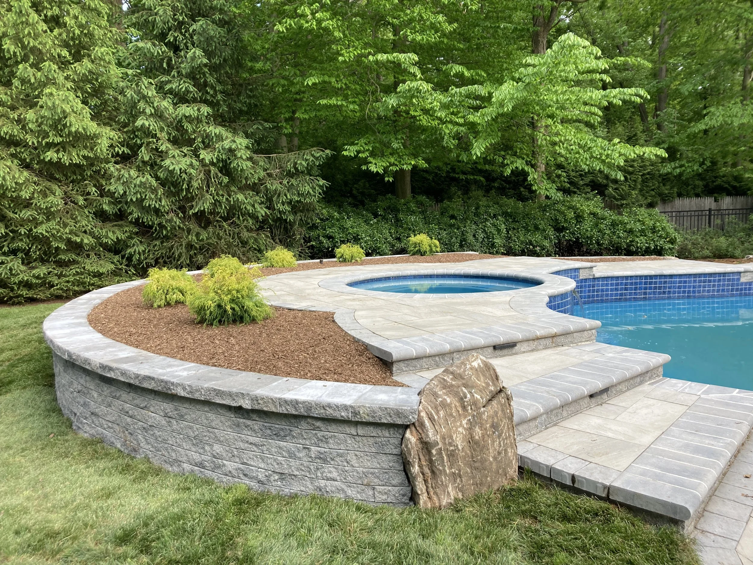 A backyard swimming pool with a hot tub, surrounded by a stone deck and landscaping, with trees and bushes in the background.